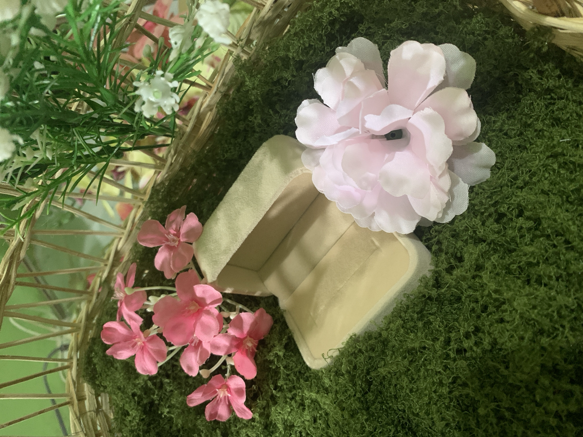 The image shows a close-up of a decorative arrangement within a woven basket. The base of the arrangement is covered in green, moss-like material. In the foreground, an open, light-colored jewelry box rests on the moss. To the left of the box are several small, pink artificial flowers. To the right and slightly behind the box is a large, pale pink artificial peony. In the upper right corner, a bunch of artificial green foliage with small white artificial flowers is visible, also contained within the basket. The background appears to be the inside of the basket and some out-of-focus elements, possibly other decorations. There are no people visible in the image, and no specific location in Kuala Pembuang, Indonesia, can be identified.