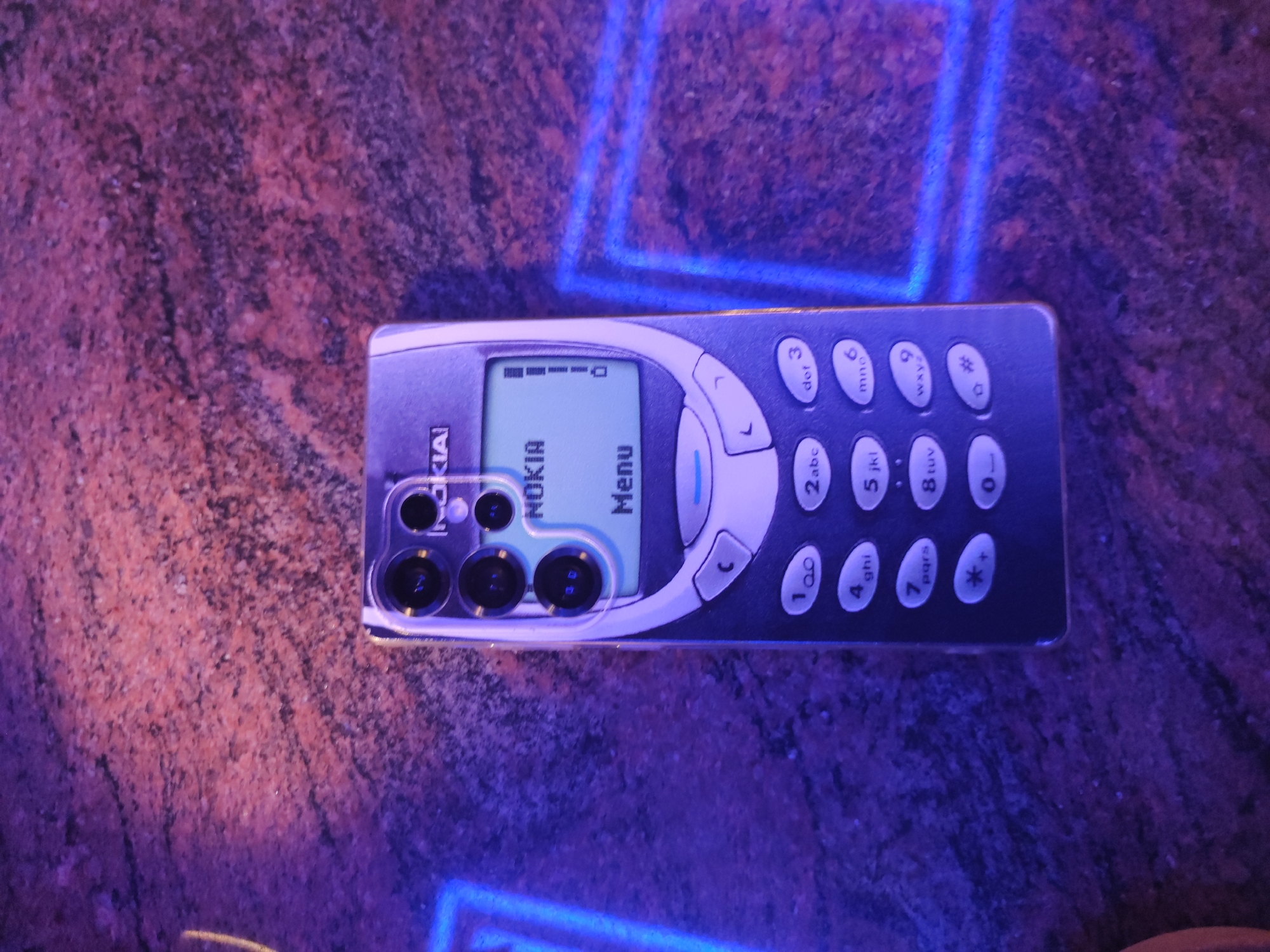 The image shows a Nokia 3310 phone lying on a speckled surface, with purple and blue lighting illuminating the scene. The phone's screen displays the Nokia logo and the word "Menu". The phone also has a modern camera array grafted on its backside, giving it a contrasting and technologically advanced look. The surface appears to be a countertop or table with a mottled pattern in reddish-brown and dark purple tones. The blue and purple lighting casts an atmospheric glow over the entire composition.