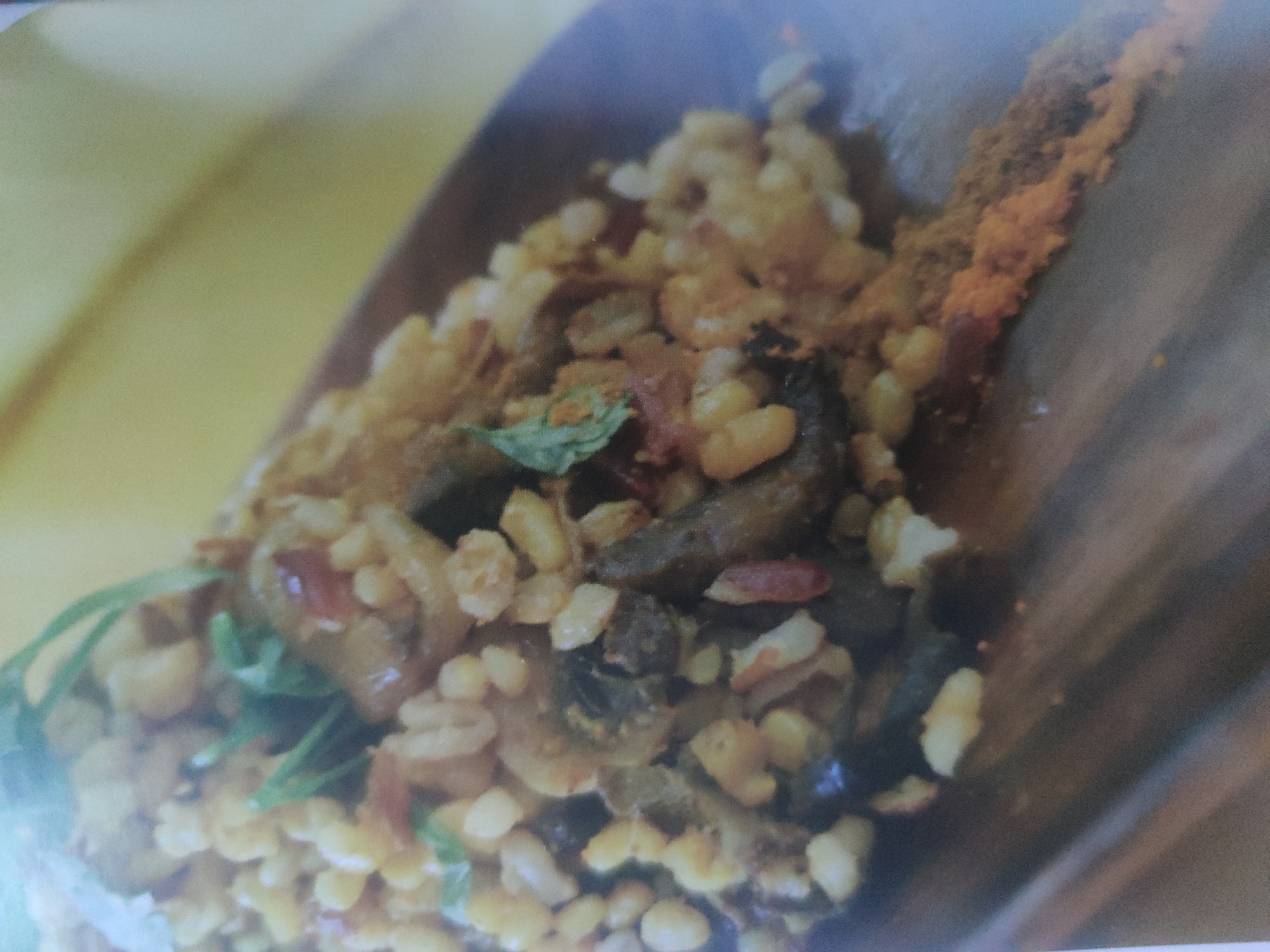 A close-up, slightly angled shot depicts a wooden bowl filled with food. The main component appears to be small, cream-colored, grain-like pieces, possibly rice or lentils, mixed with darker elements resembling vegetables or meat. Some green leafy herbs are visible on the left side. A reddish-brown powder is present along the bowl's upper edge. The setting is undefined, and no specific location or human presence is evident in the image.