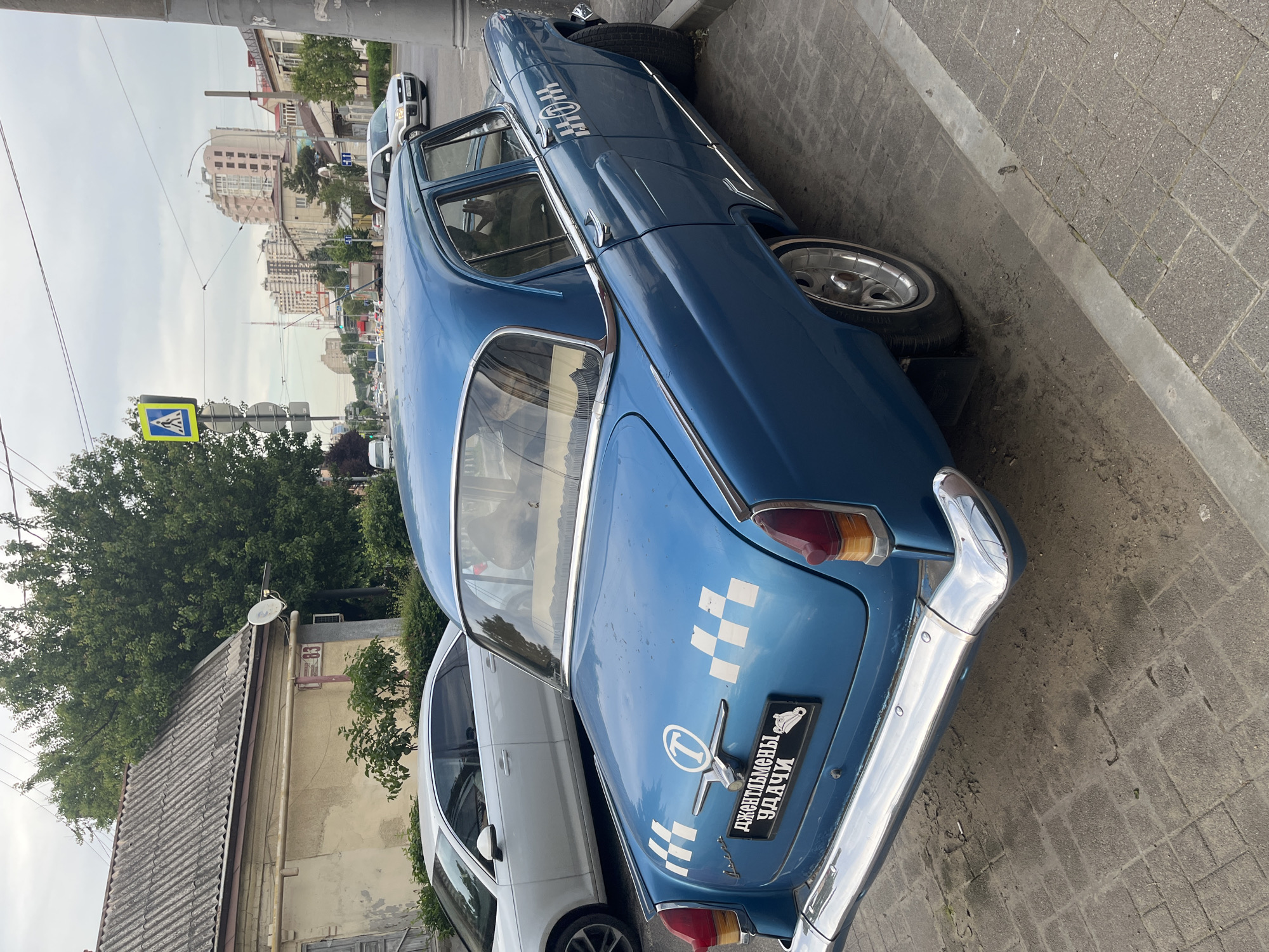 This image captures a classic blue GAZ-21 Volga sedan, customized to resemble a taxi, parked on a paved sidewalk in Krasnodar, Russia.
The scene is located at Ulitsa Postovaya, 83, Krasnodar. The number "83" is visible in red on the pale yellow stucco facade of an old, two-story building with a grey corrugated roof, situated directly behind the blue car. A satellite dish is affixed to this building.
The blue Volga features white checkerboard taxi markings on its trunk and sides, along with a circular 'T' logo on the trunk lid and near the front door handles. A decorative black license plate is affixed to the trunk, reading "ДЖЕНТЛЬМЕНЫ УДАЧИ" (Gentlemen of Fortune) in Cyrillic, accompanied by an illustration of a boot. The car has chrome bumpers, trim, and custom multi-spoke wheels with whitewall tires.
To the left of the blue Volga, a modern white sedan (possibly an Audi) is parked parallel to the curb. The pavement consists of grey rectangular tiles.
In the background, a multi-lane street is visible with light traffic, including a silver SUV (resembling a Ford Fusion/Freestyle) driving away. A prominent yellow diamond-shaped pedestrian crossing sign stands on the right side of the street. Further in the distance, modern high-rise residential buildings rise under an overcast sky. Overhead wires, characteristic of urban infrastructure, crisscross the sky. Lush green trees and bushes are scattered throughout the scene. No specific individuals are clearly visible or engaged in definable actions. The overall lighting suggests daytime conditions with diffuse light due to cloud cover.