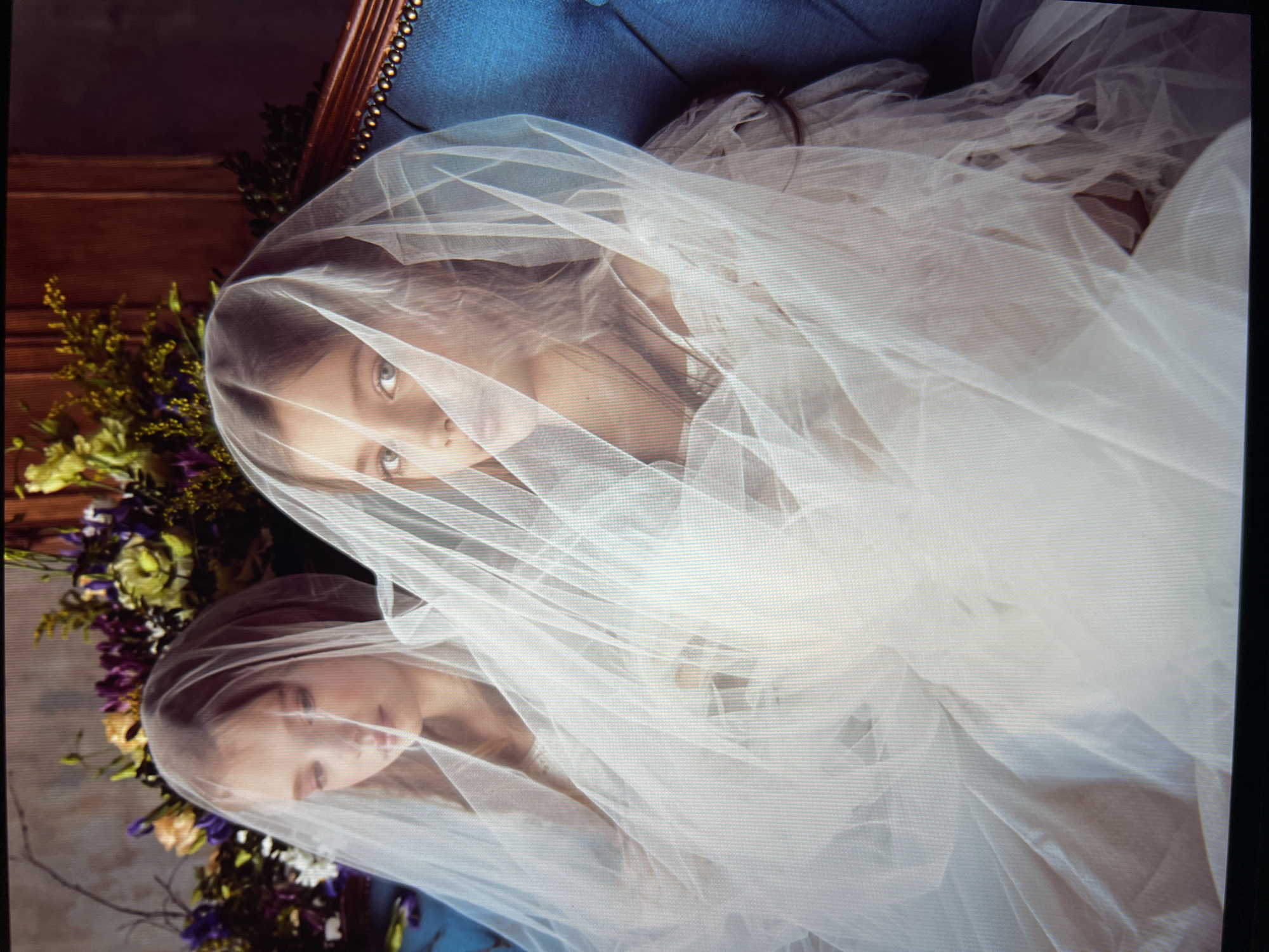 Two young girls, appearing to be twins, are seated close together on a blue tufted sofa. They are both wearing white dresses and are draped in sheer white veils that obscure most of their faces. The girl on the right is looking directly at the camera, while the girl on the left is looking down. Behind them, a lush floral arrangement featuring purple and yellow flowers is visible. The background consists of a wooden paneled wall and a dark curtain or wall. The overall aesthetic suggests a formal or artistic portrait setting. The location is unknown.