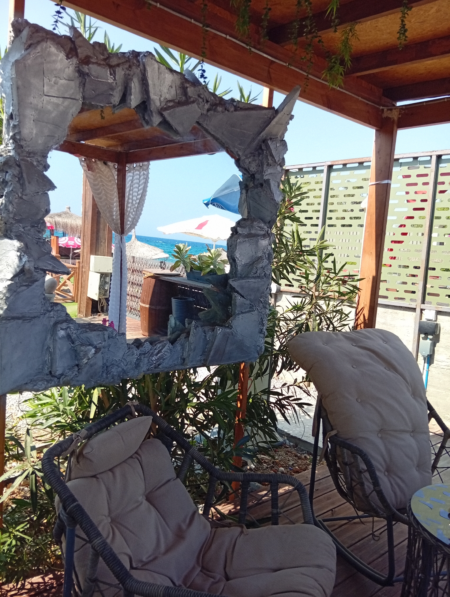 The image depicts an outdoor seating area with a view of the sea, located in Trikomo, Northern Cyprus. In the foreground, two cushioned wicker chairs are placed on a wooden deck. One chair on the left is mostly visible, with its tan cushion and woven frame. The chair on the right is partially obscured by foliage and features a large, plush tan cushion. A small, round table with a mosaic top stands to the right of the right-hand chair.
Dominating the center of the image is a large, textured, gray frame, resembling a rough stone or concrete structure, with an opening that looks out towards the sea. Behind this frame, glimpses of the beach are visible, including thatched umbrellas and lounge chairs. The sea is a clear blue, meeting a bright sky.
To the right of the frame, a green, decorative metal screen with a patterned design is visible, supported by wooden posts. Lush green plants and trees surround the seating area, adding to the tropical atmosphere. Hanging greenery is also present, trailing from the wooden ceiling structure above. The overall impression is of a relaxed, seaside establishment.