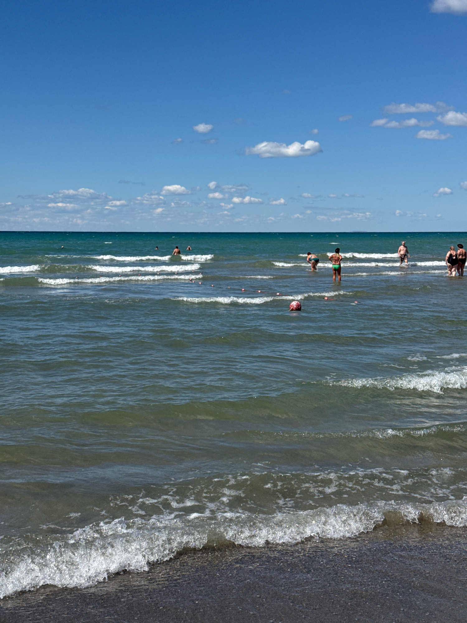 The image depicts a sunny day at a beach with a clear blue sky and scattered white clouds. The sea is a calm, greenish-blue with gentle waves rolling towards the shore. Several people are visible in the water, some wading and others swimming. In the foreground, a dark, pebbly beach is visible where the waves are breaking. A red and white ball floats in the water. In the background, a thin strip of land is visible on the horizon, possibly another coastline. The overall scene is one of leisure and recreation on a summer day. The location is identified as the city of Anapa, Russia.