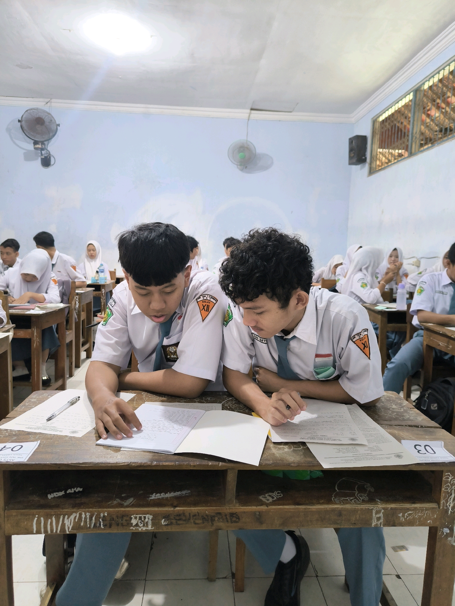 The image depicts a classroom setting, likely a school, in Kebumen, Indonesia. Two young male students, wearing white school uniforms with green and orange insignias, are seated at wooden desks in the foreground. The student on the left is looking down at a notebook and papers, holding a pen, while the student on the right is leaning forward, also focused on papers on his desk. Their uniforms suggest they are in secondary school, likely around 11th or 12th grade, given the roman numeral XII on the insignia of the student on the left.
The classroom environment appears somewhat dated, with light blue walls and a white ceiling with fluorescent lighting. Two oscillating fans are mounted on the walls, indicating a warm climate. A barred window is visible on the right wall, showing some indistinct shapes beyond. Other students in similar uniforms are visible in the background, also engaged in individual work at their desks. The desks themselves are made of wood and show signs of wear and graffiti. Papers, pens, and notebooks are scattered across the desks. A black backpack is visible on the floor next to the desk of the student on the right. The overall impression is one of a focused academic activity, possibly an examination or a study session, within a typical Indonesian school classroom.