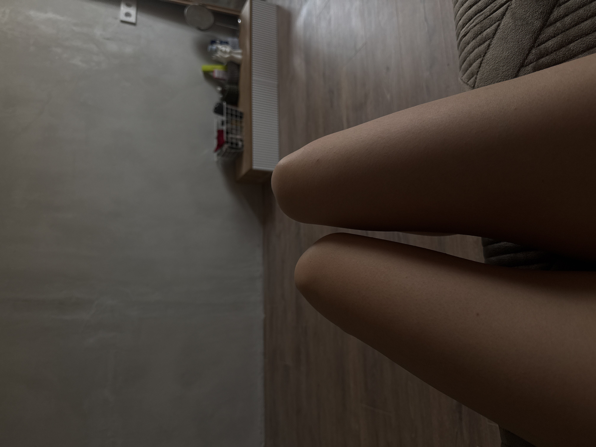 The image depicts two human legs in close proximity to an upholstered surface. The legs appear to be resting on the upholstery. In the background, a wall and a wooden floor are visible, along with a shelving unit containing various items. The location of Votkinsk, Russia, cannot be determined from the image.