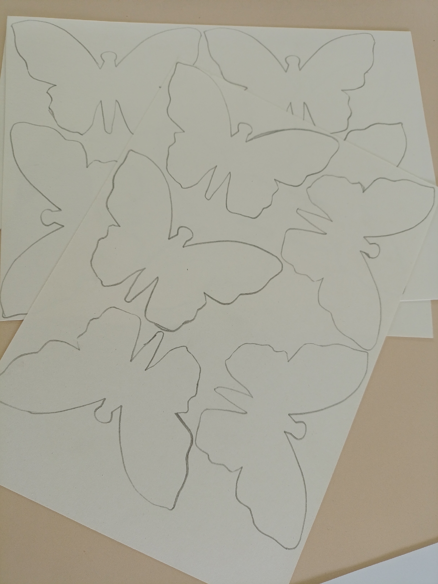 The image shows several sheets of white paper with butterfly outlines drawn on them in pencil. The sheets are stacked on top of each other, slightly askew, revealing portions of each butterfly drawing. The background is a plain, light brown surface. The image does not contain any information to suggest a specific location or a connection to the city of Chaikovsky, Russia.