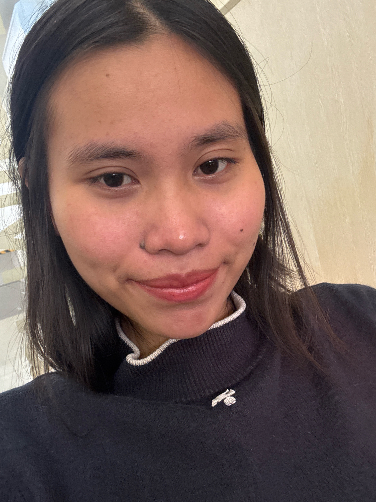 A young woman with long, dark hair and fair skin is looking directly at the camera. She is wearing a dark blue, high-necked sweater with a white and light grey ribbed trim around the neckline. A small, silver-colored pin with a geometric design is attached to the left side of her sweater. Her makeup includes lip gloss and a slight blush on her cheeks. Her background appears to be an indoor setting with a textured wall. There is no discernible indication of location within the city of East Jakarta, Indonesia.