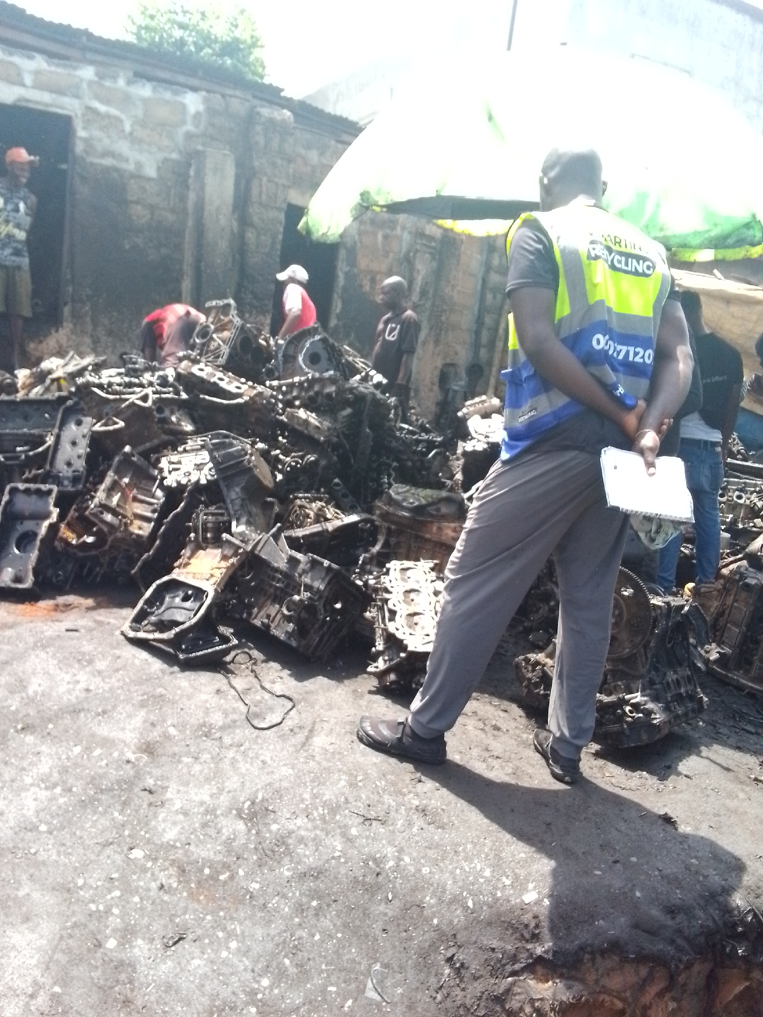 The image depicts a recycling operation in Onitsha, Nigeria. A large pile of what appear to be used car engine parts dominates the scene. Several people are around the engine parts, some of whom seem to be sorting or examining them. One man stands in the foreground wearing a reflective vest marked with the word "Recycling" and a phone number. He holds a small notebook in his hands. The ground appears oily and stained. In the background, there are simple, older buildings and a makeshift awning.
