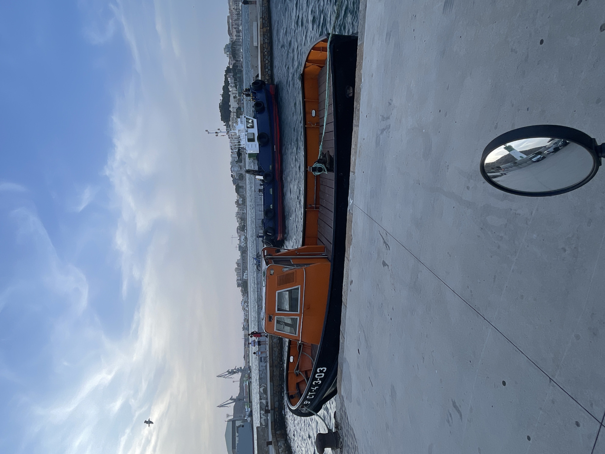 An orange and black tugboat, bearing the identification number 5 CT-43-03, is moored to a concrete quay. The boat has a wooden deck and a cabin structure with windows. Behind it, a larger blue and white tugboat is also in the water. In the background, the city of Cartagena, Spain, is visible with its buildings and a hill. Cranes suggest an industrial or port area. The sky is partly cloudy with a bird flying in the upper left quadrant. A circular mirror, likely from a vehicle, reflects buildings and a street scene in the lower right corner of the image. The overall scene depicts a waterfront area in a Spanish port.