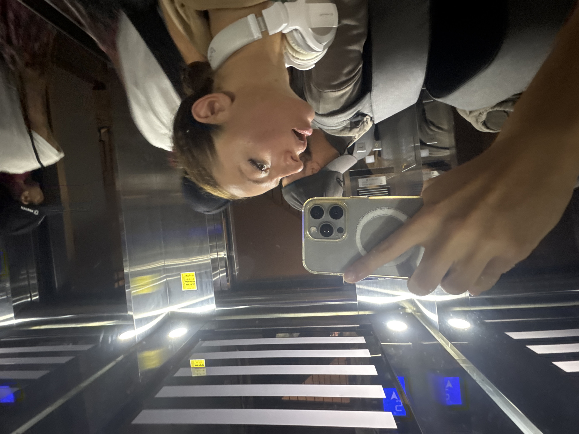 A woman with headphones is taking a selfie in an elevator with a metal interior. The elevator appears to have horizontally striped flooring. The reflection in the elevator shows another person in the background. Blue up arrow and down arrow signs are visible. There is insufficient information to determine if this image was taken in Đà Nẵng, Vietnam.