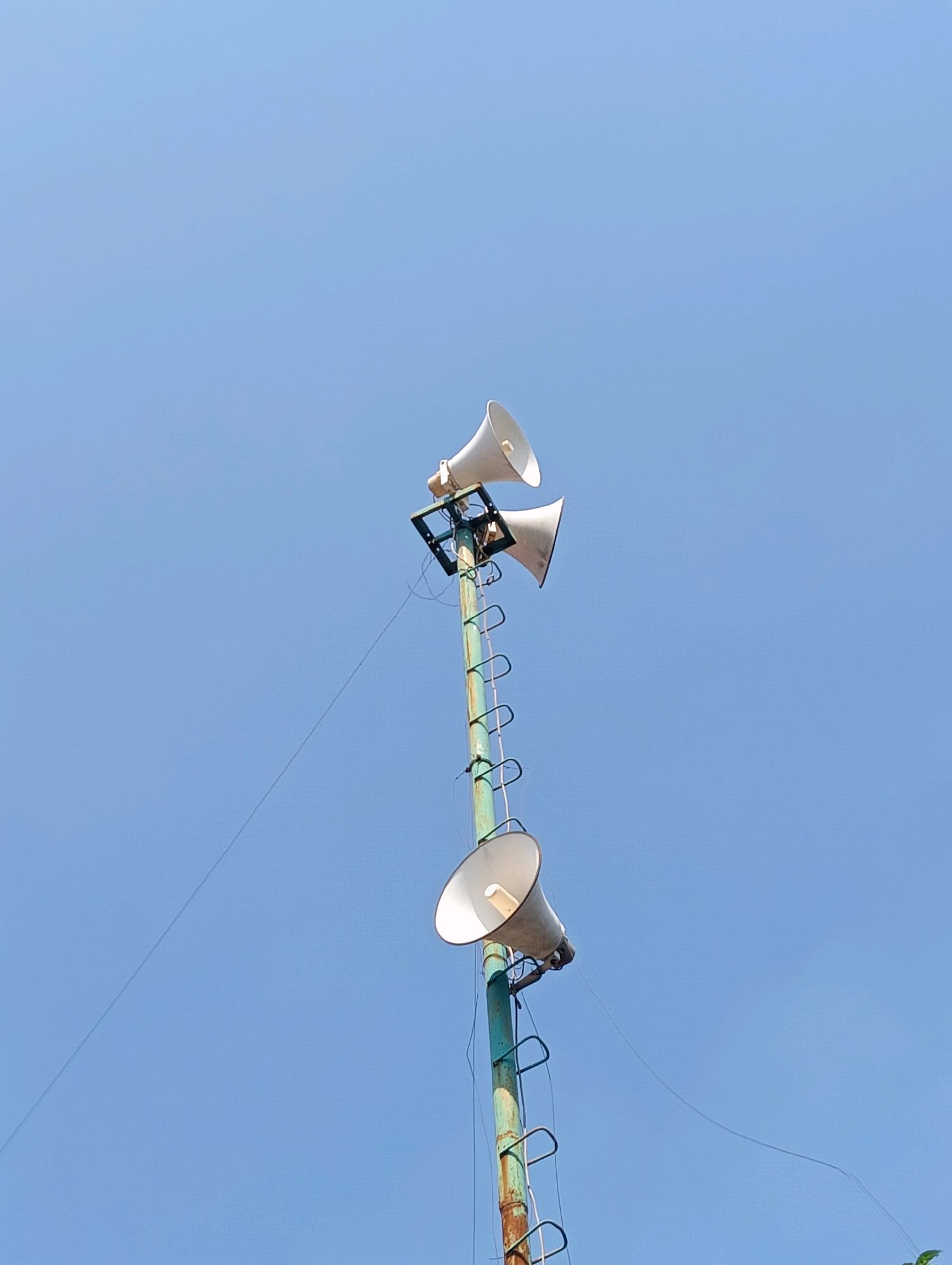 A weathered, green metal pole stands tall against a clear blue sky in Kecamatan Pati, Indonesia, supporting three white loudspeakers arranged vertically. The pole shows signs of rust and age. A ladder, also made of metal, is attached to the pole, extending upwards alongside the loudspeakers. Thin wires are connected to the speakers and run down the pole and across the sky. The lower part of the pole is partially obscured by a hint of green foliage at the very bottom of the frame. The scene is bathed in bright daylight, casting no visible shadows.