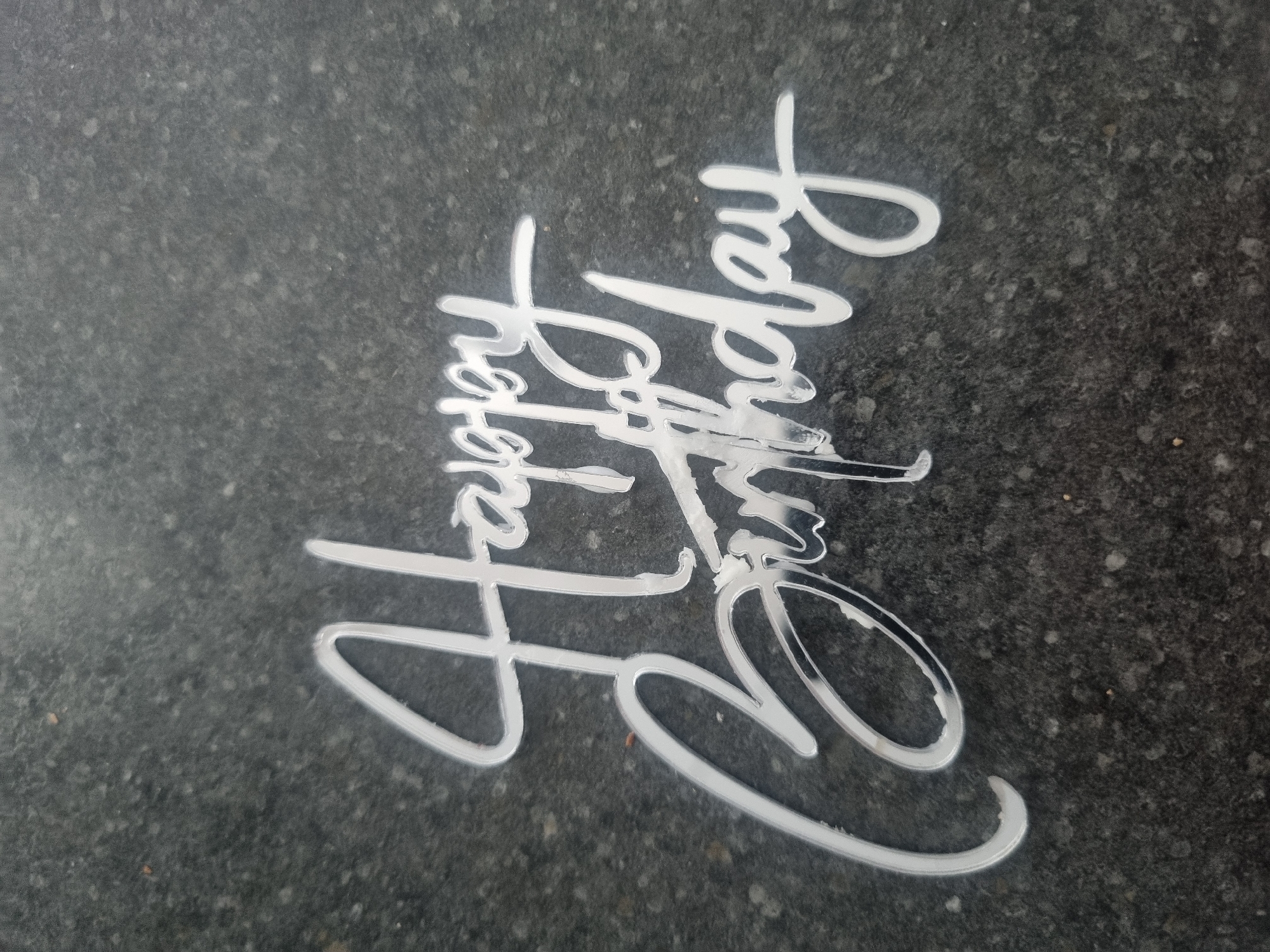 A close-up, overhead shot shows a white, reflective acrylic cake topper that reads "Happy Birthday." The lettering is in a cursive font, with "Happy" positioned above and slightly to the left of "Birthday." The cake topper is placed on a dark, speckled surface that appears to be granite or a similar material. There are no people or any other objects visible in the frame. The lighting is moderate, highlighting the reflective nature of the cake topper. The location is not specified within the image itself.