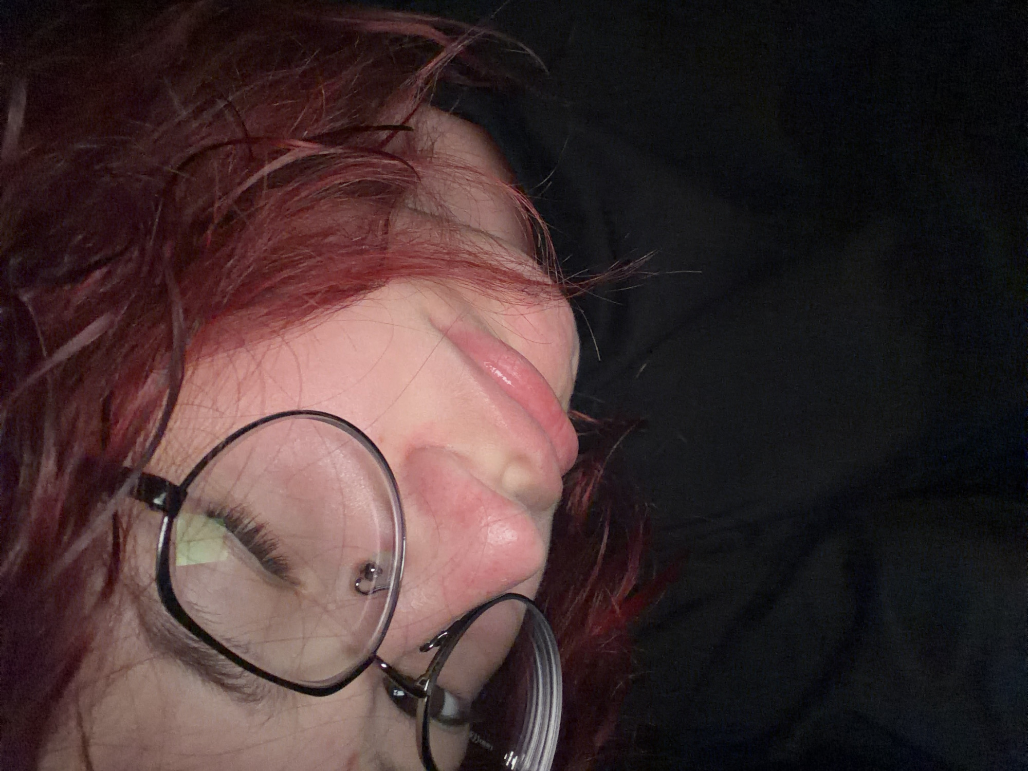 The image is a close-up, downward-angle shot of a person's face, focusing on their eye, nose, and mouth. They are wearing round-rimmed glasses with black frames. Their hair is a reddish-brown color and appears somewhat messy, falling across their face and partially obscuring their glasses. The person's skin tone is fair, and their lips are slightly parted and appear to be glossy. The background is predominantly dark, with hints of clothing, possibly a black shirt or jacket. The lighting is dim, casting shadows and highlighting certain features. There is no visible indication of the specific location within Chelyabinsk, Russia, or any activity taking place.