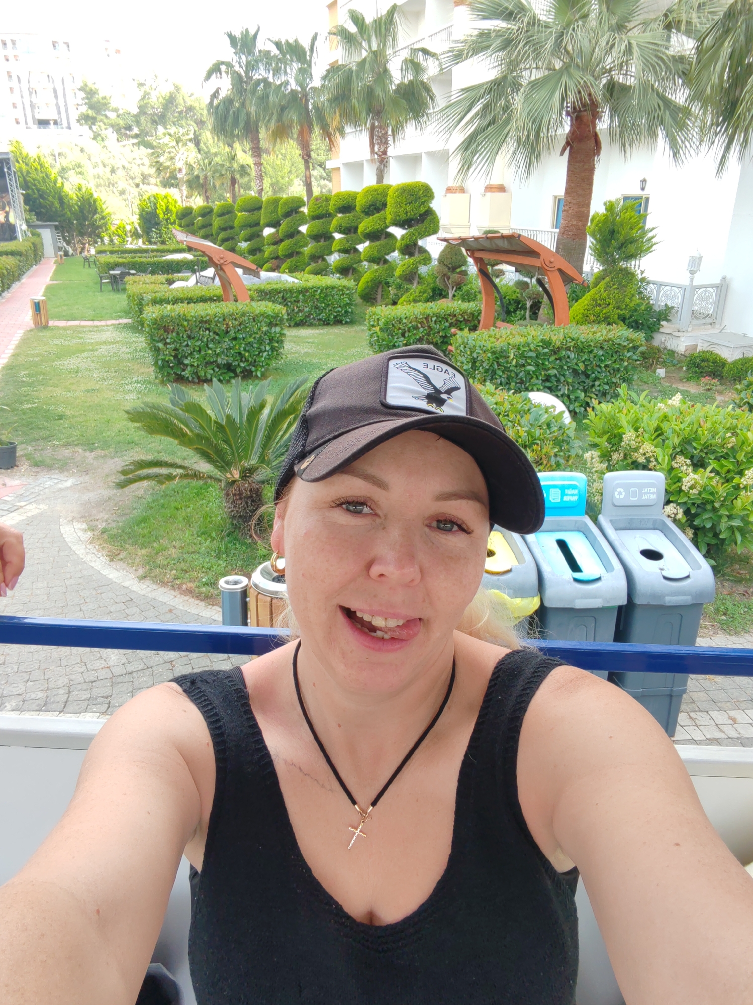 A medium shot shows a fair-skinned woman in a black tank top and black baseball cap with an eagle emblem, taking a selfie with a playful expression; she is sticking her tongue out. She is standing in front of three recycling bins, each labeled for specific materials in a foreign language.
Behind her is a landscaped garden with various bushes, trees, and palm trees. Decorative benches can be seen among the greenery. The location is Alanya, Turkey, as indicated by the presence of specific landscaping styles commonly found in the city. A building with white exterior walls and decorative architecture is partially visible in the background. The overall setting suggests a tourist area or resort location. A small part of another person's arm is visible at the left side of the frame.