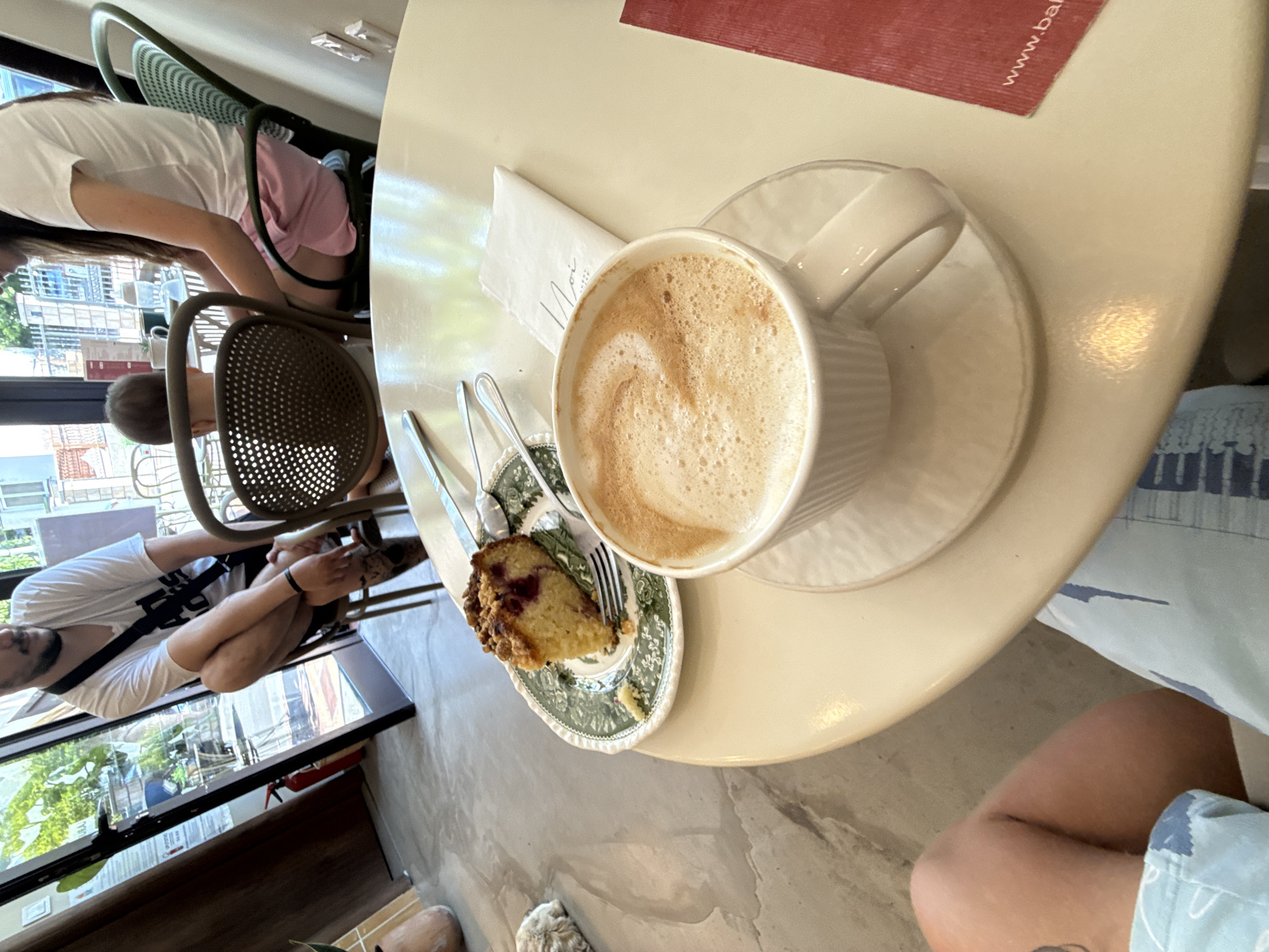 The image shows a close-up view of a table at an outdoor cafe, possibly in Budva, Montenegro, as evidenced by a red placemat with the partial text "www.ba...". In the foreground, a white cup of frothy cappuccino sits on a saucer, with a piece of cake on a decorative green and white plate to its left. A fork and spoon are also present on the plate. In the background, a man in a white t-shirt and shorts is partially visible, sitting on a chair and looking away from the camera. To his right, a woman in a white t-shirt and pink shorts is seated. A small child is also visible between the man and the woman, sitting in a chair. The cafe appears to have large glass doors and windows, with greenery visible outside. The table is round and cream-colored, and the floor is a light-colored polished concrete.
