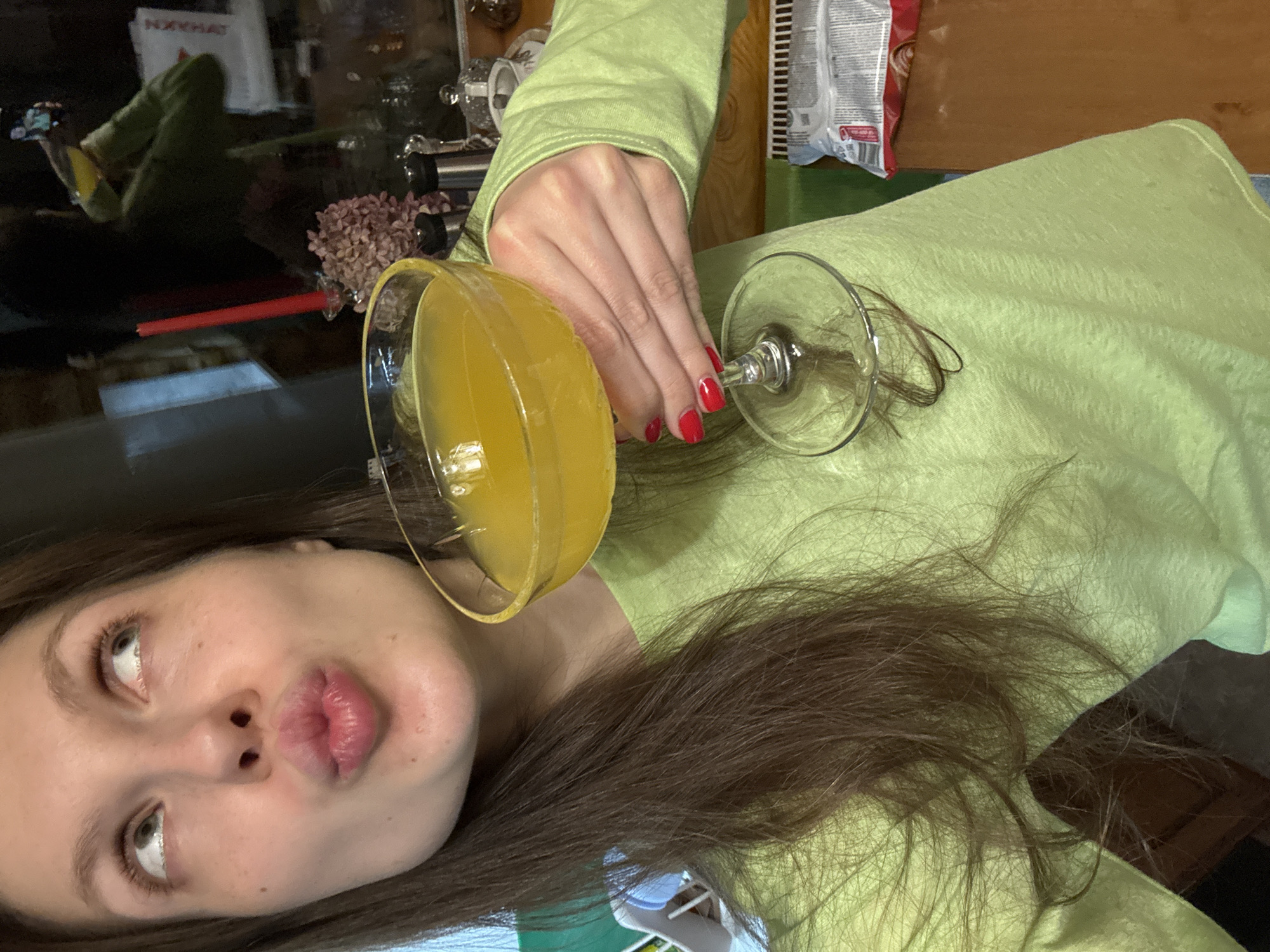 The image shows a woman with brown hair and a light green long-sleeved shirt holding a glass of yellow liquid. She is looking at the camera with pursed lips. The fingernails on her hand are painted red. An overturned empty glass with strands of hair is lying on the shirt near her hand. The background is cluttered with various items including a table, a bag of chips, and what appears to be a floral arrangement. No information in the image identifies the location as Tomilino, Russia.