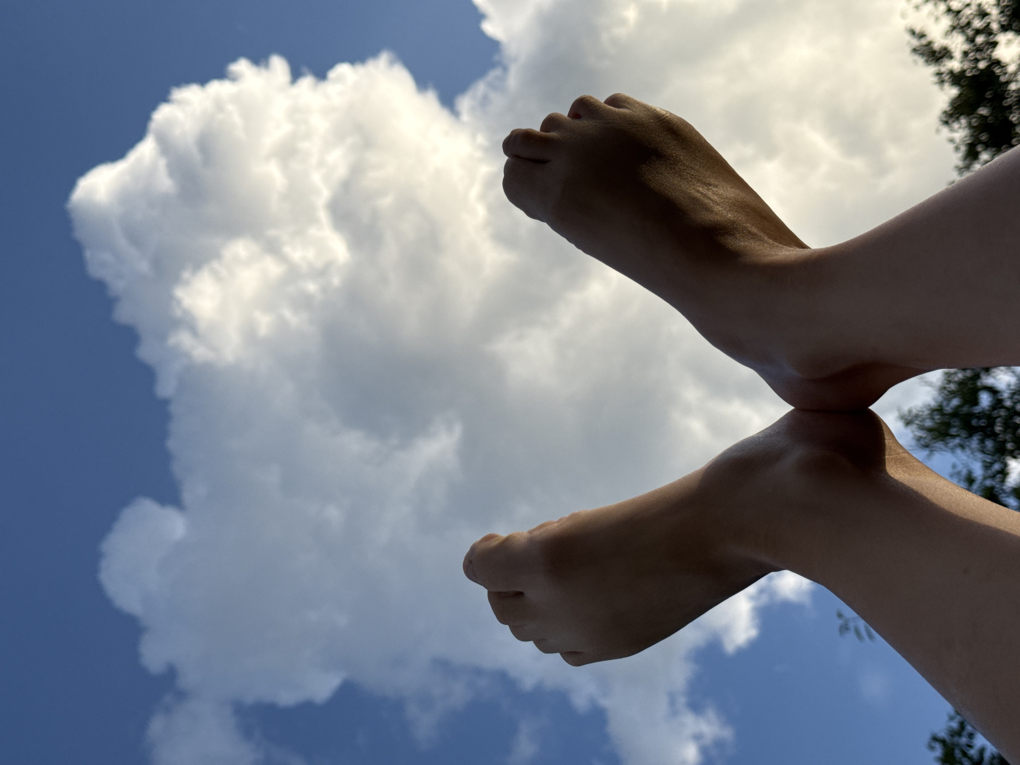 Two bare feet are positioned in an 'X' shape against a blue sky with large, white cumulus clouds. The feet are illuminated by sunlight from the side, creating shadows on their undersides. The background also shows blurry green foliage at the bottom of the frame, suggesting trees or bushes. The image appears to be taken outdoors, possibly in a park or open area. The location in Vetchluzhsky, Russia, is not visually evident from the image itself.