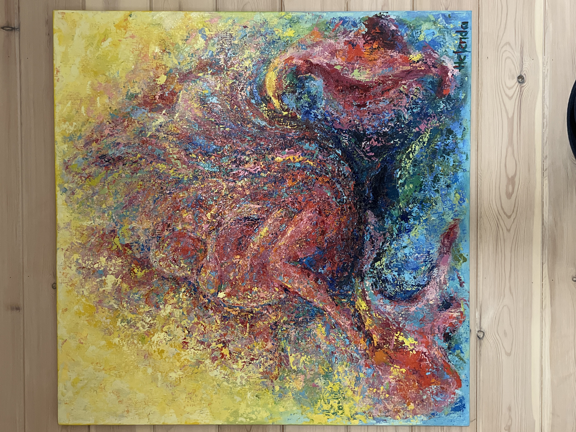 The image displays an abstract painting with vibrant colors, primarily featuring reds, yellows, blues, and purples. The style is heavily textured, with thick brushstrokes that create a sense of movement and energy. The painting appears to depict two figures, possibly intertwined or in a state of embrace, rendered in an abstract, impressionistic manner. The background is a wash of yellow and lighter hues, contrasting with the intense colors of the figures. The artwork is signed in the lower right corner with "Helenda". The painting is hung on a wooden paneled wall, suggesting an indoor setting. The location in Angarskie Khutora, Russia is not discernible from the image itself.
