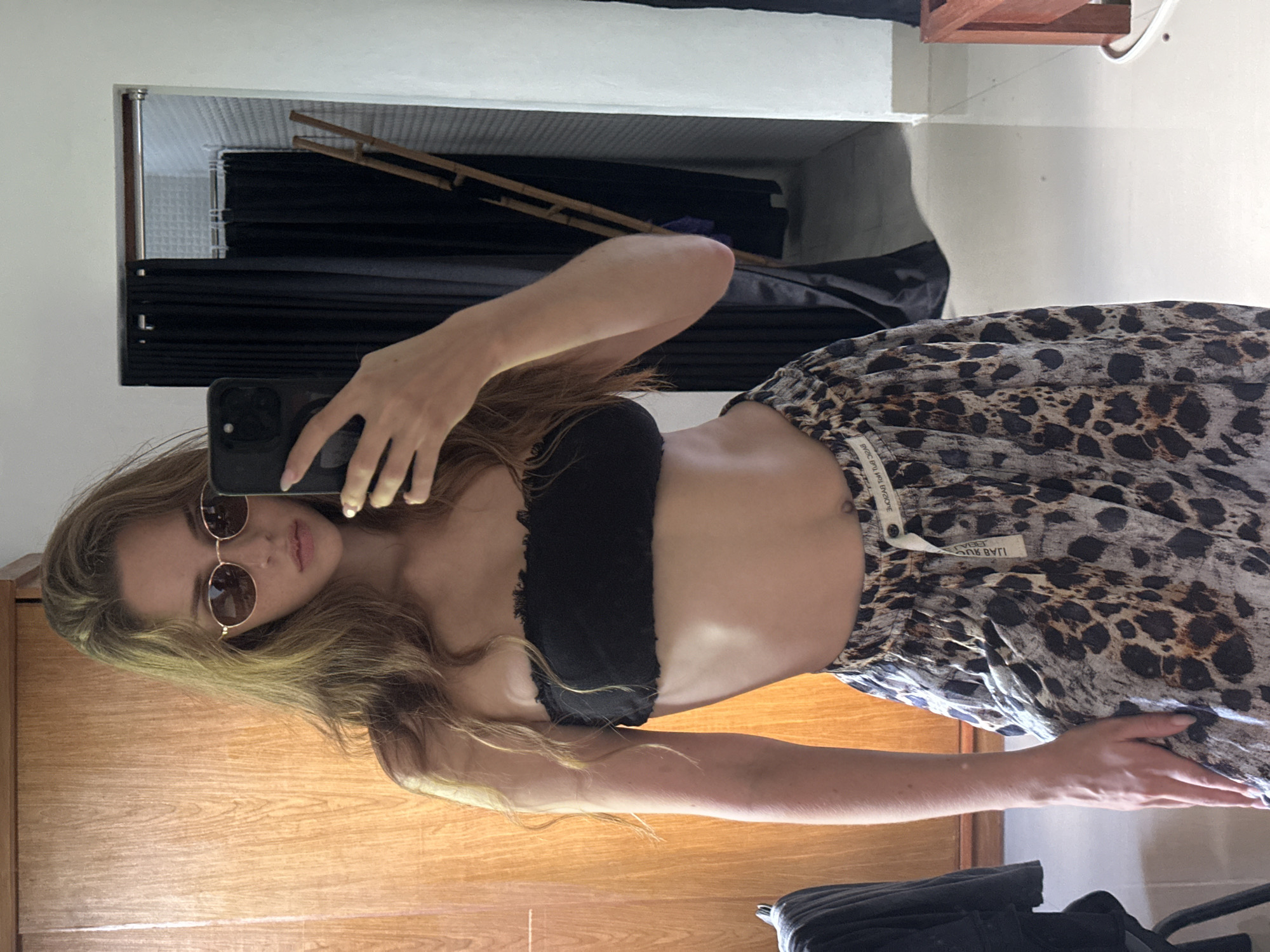 The image displays a young female taking a mirror selfie indoors. The subject has long, wavy brown hair and wears small, round, gold-rimmed sunglasses with dark lenses. She is dressed in a black strapless bandeau top and loose-fitting, leopard-print pants or a skirt featuring a white drawstring and a partially visible inverted label that reads "BASIC BUT HOT". Her midriff is exposed. She holds a dark-colored smartphone in her right hand, angled towards the mirror to capture the reflection. Her fingernails are painted a light, possibly metallic, color.
The background, observed through the mirror's reflection, includes a large, light brown wooden wardrobe or closet on the left side of the frame. On the right, a dark curtain partially screens an inner area revealing light-colored, possibly tiled, walls and a wooden ladder leaning against a far wall. Another black curtain is visible deeper within this screened space. The floor is covered with light, reflective tiles. A fragment of a reddish-brown wooden object, possibly furniture, is present in the lower right corner. The environment appears to be an indoor space, such as a changing room or bedroom, situated in North Kuta, Indonesia, as specified.