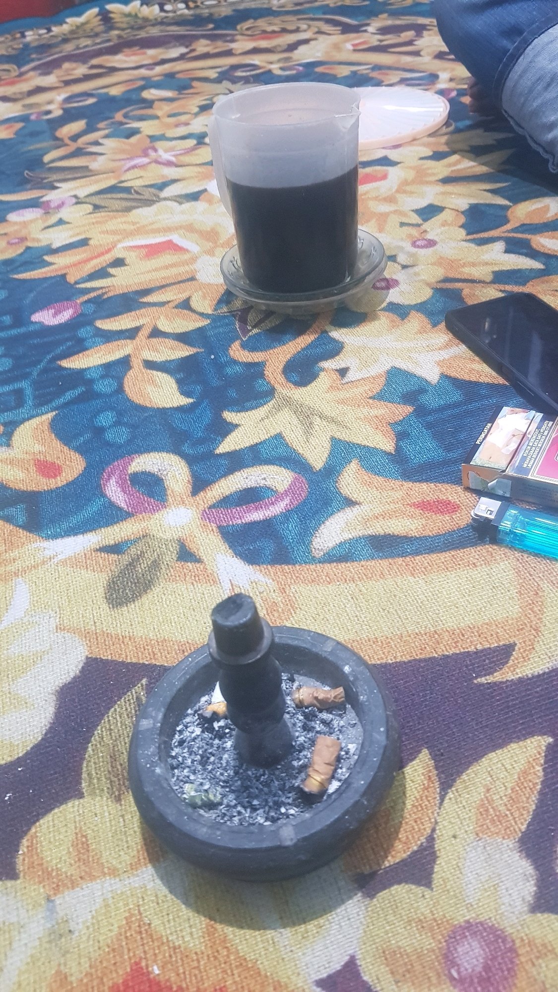 The image depicts a close-up view of a patterned tablecloth, likely in Bakalan, Indonesia. On the tablecloth, there is a clear plastic cup filled with a dark liquid, presumably coffee, placed on a small glass saucer. To the right of the cup, a pack of cigarettes and a blue lighter are visible. In the foreground, a stone ashtray contains cigarette butts and ash. A person's leg, clad in blue jeans, is partially visible in the upper right corner of the frame, suggesting the presence of an individual. The overall scene suggests a moment of relaxation or a break.