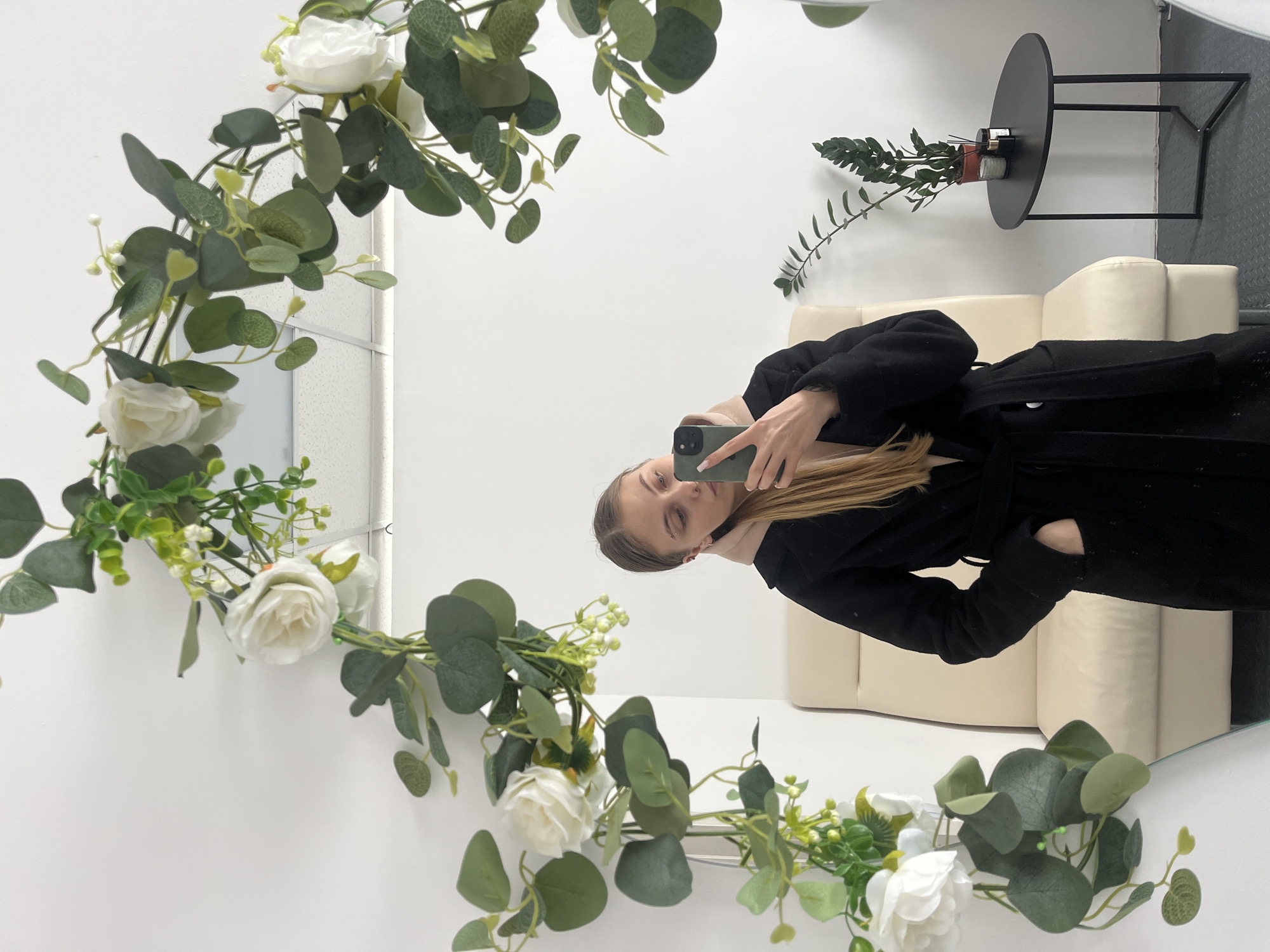 A young female subject is observed taking a mirror selfie within an indoor location in the city of Фоки, Russia. The subject possesses long, straight, light brown hair and is dressed in a black, belted wrap coat over a lighter-colored garment visible at the neckline. Her left hand is inserted into the coat's pocket. She holds a dark green smartphone in her right hand, angled to capture her reflection. An eyebrow piercing is visible on her left eyebrow. The mirror, which is the primary reflective surface, is framed by an artificial garland composed of white roses, green eucalyptus-like leaves, and other small green foliage. The reflection in the mirror reveals a room with white walls. To the subject's left, a cream-colored upholstered armchair or recliner is visible. To her right, a black oval side table holds a potted green plant, two dark-colored candles, and a smaller decorative object. The floor is dark in tone, and a white paneled ceiling is reflected along the upper edge of the mirror.