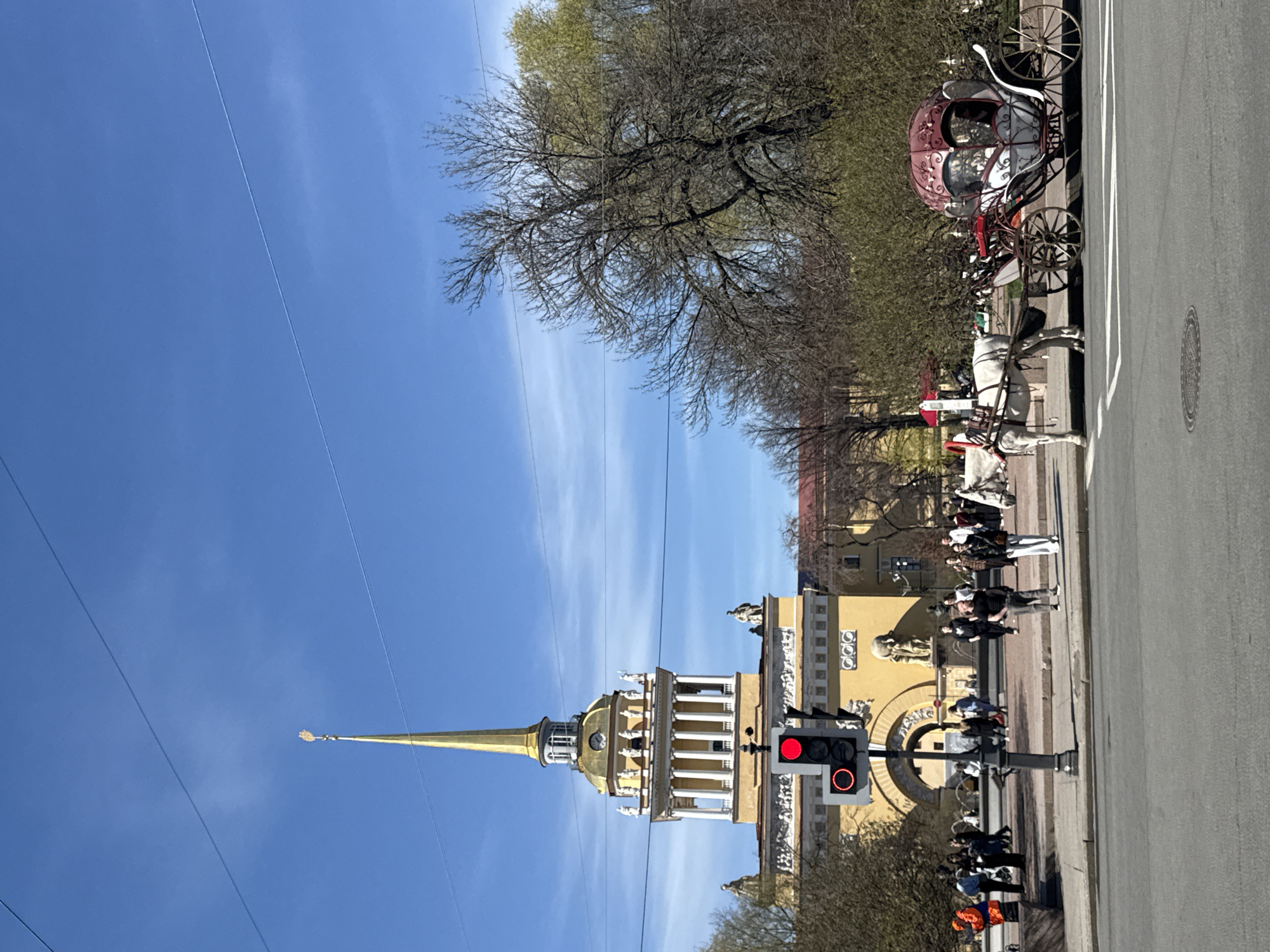 The image depicts a sunny day in Saint Petersburg, Russia, with the Admiralty building prominently visible on the left side of the frame. The golden spire of the Admiralty stands out against the clear blue sky with scattered wisps of clouds. A horse-drawn carriage, complete with a red and white ornate carriage, is positioned on the right side of the street, being pulled by a white horse. Several people are standing on the sidewalk near a traffic light, which is displaying a red light. The street appears to be paved and relatively wide. Trees line the background behind the Admiralty and along the street where the carriage is located. Power lines run across the sky.