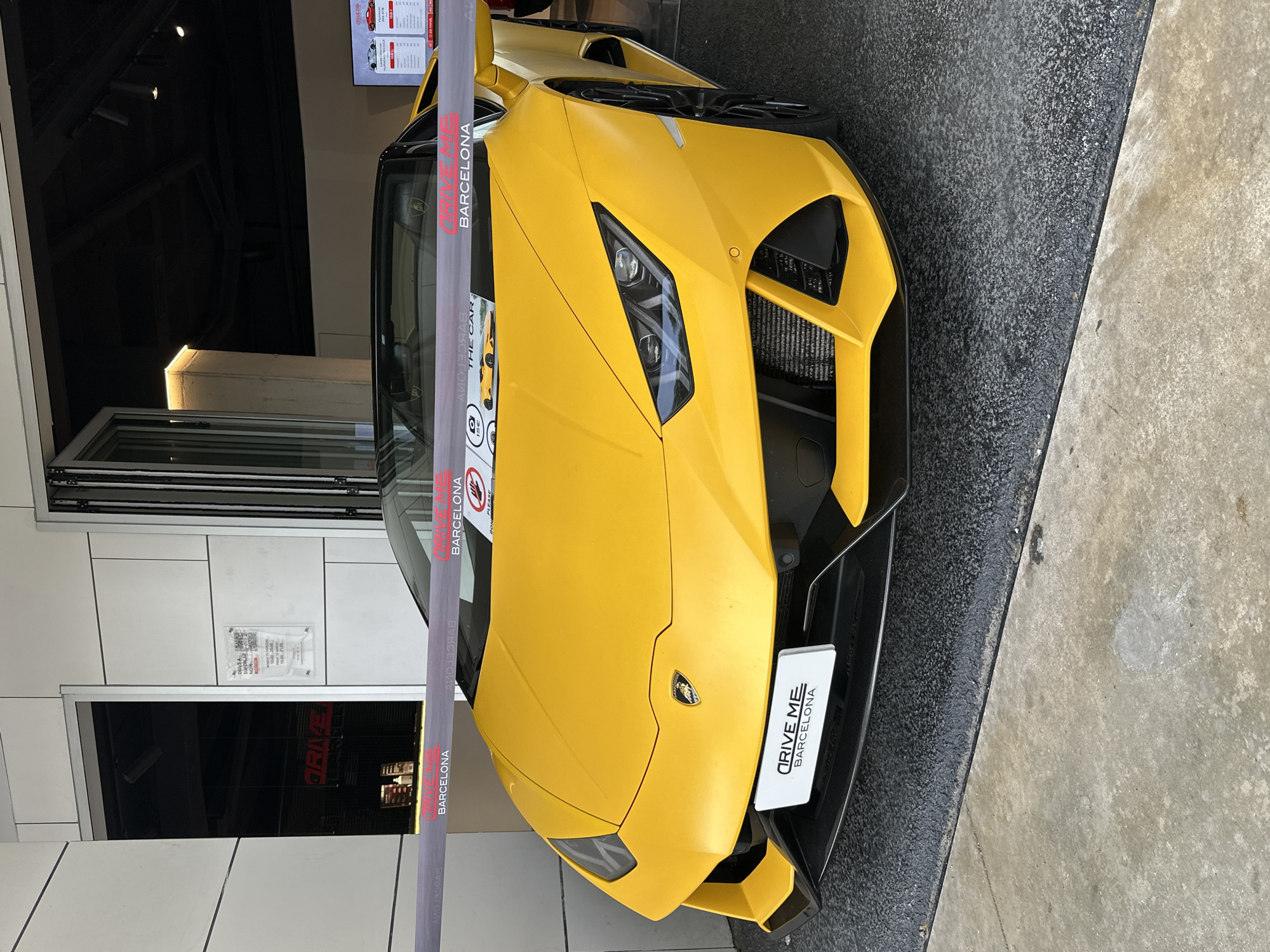 The image displays a bright yellow Lamborghini Huracan convertible supercar prominently positioned inside a commercial establishment in Barcelona, Spain. The vehicle, facing slightly towards the left, occupies the entrance area, situated on a dark, granular flooring. A grey and red barrier tape, prominently featuring the text "DRIVE ME BARCELONA" in white, stretches horizontally across the car's front at bumper height. The tape also includes a "no hands" icon and a small informational placard below, which reads "THE CAR" and "15€" accompanied by a camera icon.
The Lamborghini exhibits its characteristic front design, including the emblem on the hood, intricate LED headlights, and large, angular air intakes. A white rectangular sign on the lower front grille also bears the "DRIVE ME BARCELONA" branding.
The immediate background reveals the building's exterior, composed of light-toned, rectilinear wall panels and a large glass folding door system that is partially open. Inside the dimly lit interior, a digital display screen is visible on the right, showcasing images of supercars and associated pricing. A clear acrylic sign affixed to a wall panel on the right explicitly advertises "DRIVE A SUPERCAR BARCELONA," detailing options for a "LAMBORGHINI HURACAN / FERRARI" with listed prices and operating hours: "MONDAY TO THURSDAY 10:00 - 21:00" and "FRIDAY TO SUNDAY 10:00 - 22:00." No individuals are present within the frame. The foreground consists of a worn, grey concrete surface. The cumulative textual evidence confirms the location as Barcelona and identifies the establishment as a supercar driving experience business.