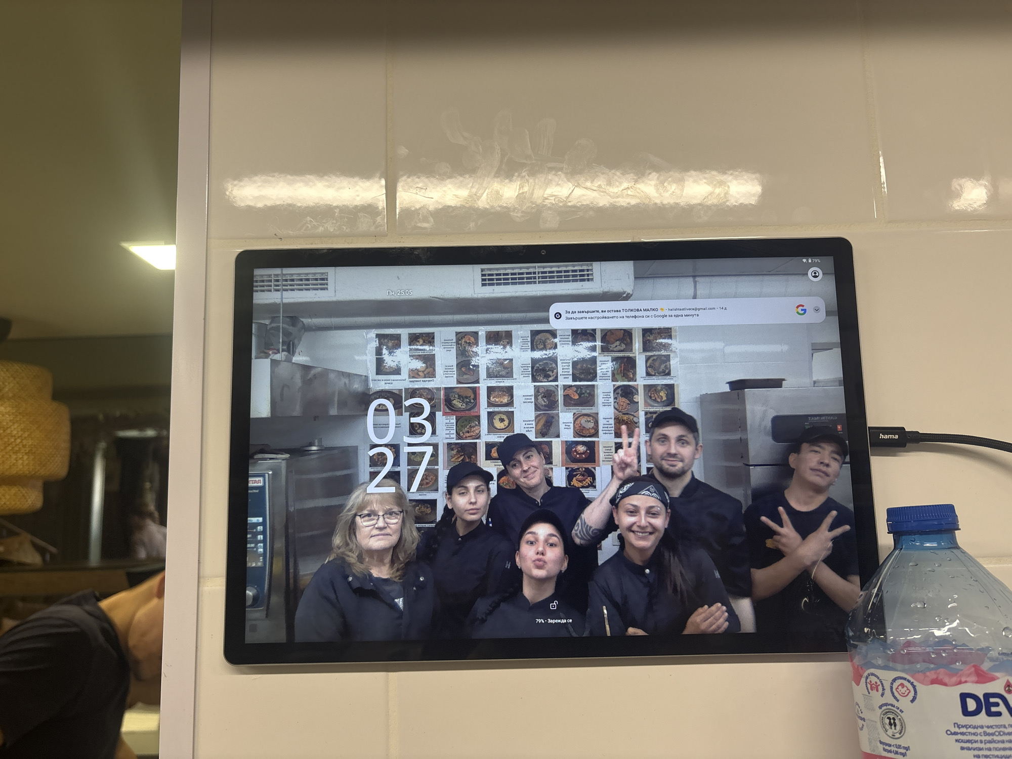 The image depicts a tablet, likely mounted on a wall in what appears to be a kitchen environment, possibly in Veliko Tarnovo, Bulgaria. The tablet displays a photo of a group of seven individuals, presumably kitchen staff, posing in front of a menu board. The photo includes the date "03 27" overlaid on the image. The tablet's screen also shows parts of a Chrome browser with search bar in Bulgarian, a Google prompt in Bulgarian, and a battery indicator showing 79%. To the right of the tablet is a bottle of "Devin" brand water, which is a Bulgarian brand. The tablet is connected to a cable, labeled "hama." The wall is covered with white, possibly ceramic, tiles.
