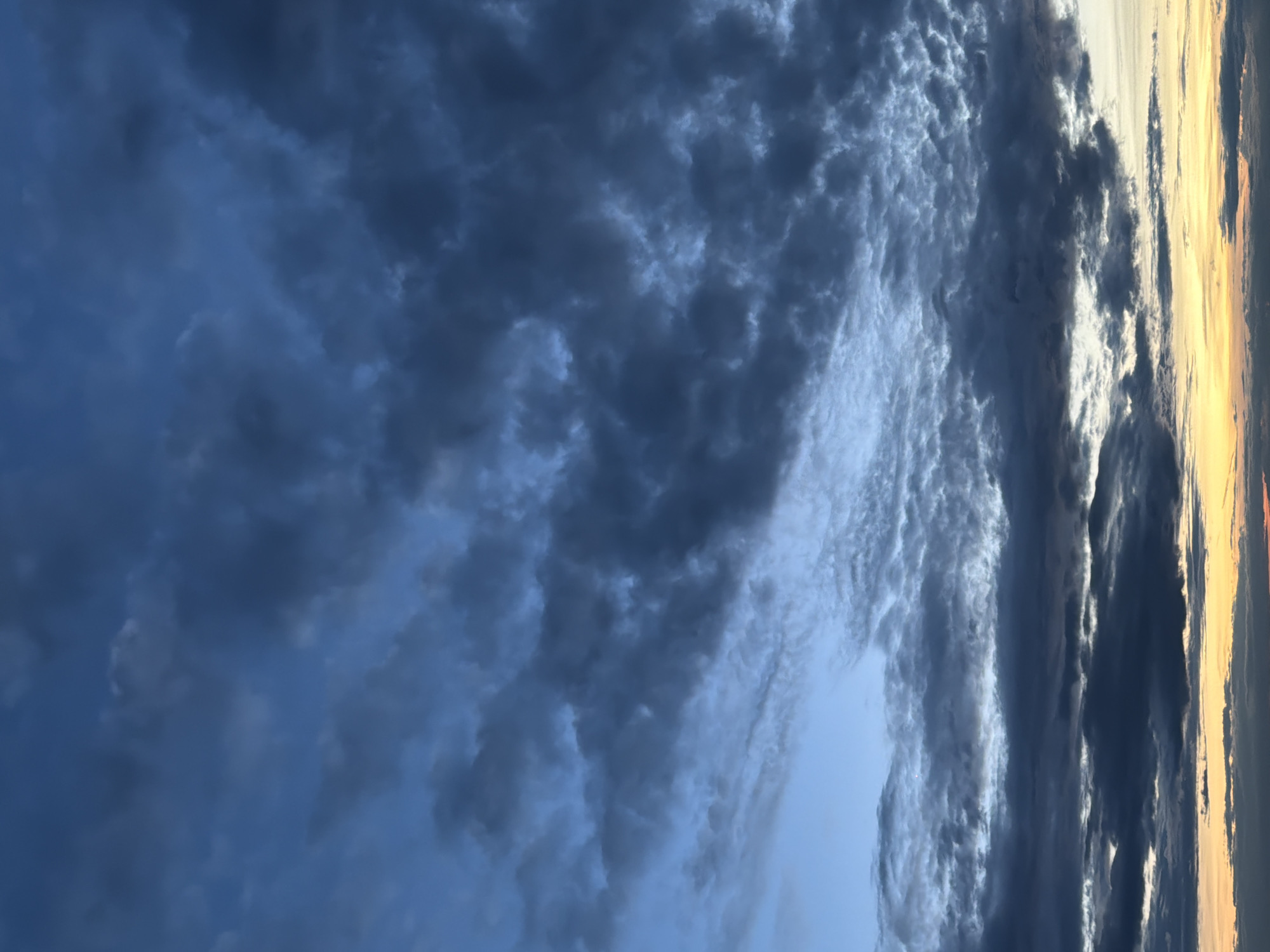 The image shows a sky filled with a variety of cloud formations. On the left side, dark, dense clouds dominate, creating a stormy or overcast appearance. Towards the center and right, the clouds become lighter and more textured, with some areas appearing wispy. Along the far right edge, there is a hint of orange and yellow hues, suggesting either sunrise or sunset. The overall composition gives a sense of atmospheric depth and drama. The location and city are unspecified.