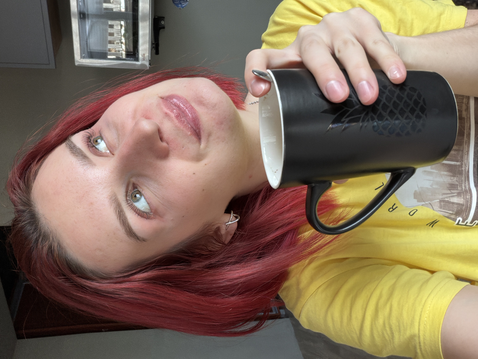 A highly tilted, close-up indoor portrait captures a person, likely female, with vibrant red hair, fair skin, and light green or hazel eyes. The image is set in Chaikovsky, Russia. The subject is wearing a bright yellow t-shirt, which has a partially visible graphic design or text on it. She holds a black ceramic mug with an embossed pineapple pattern in her right hand, positioned near her face. The mug appears to be mostly empty or contains a light-colored liquid.
The person looks upwards with a subtle, relaxed smile, her lips appearing glossy. Notable details include a silver ear cuff piercing on her left ear and well-defined eyelashes and eyebrows. The background shows a light gray wall or ceiling and a rectangular, silver-toned appliance or heater, possibly a "Volo" brand as indicated by visible text. This appliance reflects bright, possibly natural, light, suggesting it is daytime. The overall lighting is soft and even.