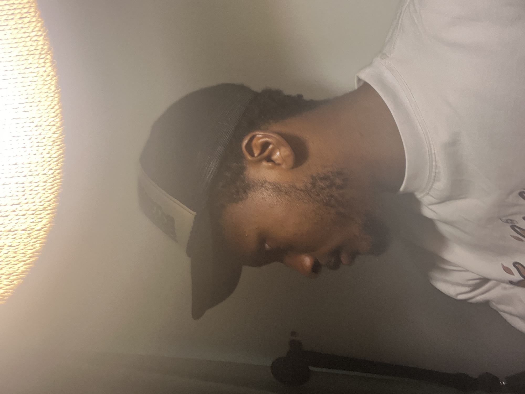 The image captures a serene, close-up profile of a person resting indoors. The subject, a male with dark skin, is depicted from the right side, with his head tilted as if lying down or leaning against a surface. He wears a dark baseball cap, possibly a trucker style, with a light-colored front panel that might bear an illegible logo or text. His eyes are closed or cast downwards, conveying a sense of peace, sleep, or deep thought. He has short, dark hair and a neatly trimmed beard or stubble.
He is dressed in a white, short-sleeved top, likely a t-shirt or sweatshirt, which shows faint, indistinct patterns or text in a darker shade on the chest area. A striking element of the scene is the bright, warm light source positioned on the left side of the frame. This light appears to shine through a textured, woven material, possibly a lamp shade or a window curtain, casting a soft, hazy glow that bathes the scene in warmth and diffusion.
The background consists primarily of a plain, light-colored wall, creating a simple and uncluttered setting. Towards the bottom right of the image, a dark, ambiguous object, possibly a stand or a piece of furniture, is partially visible. The overall mood of the image is tranquil and contemplative, enhanced by the soft lighting and the subject's relaxed posture. The location context provided is Gwarinpa, Nigeria, though the image itself does not contain specific visual cues to this location. No clearly legible text is visible within the image.