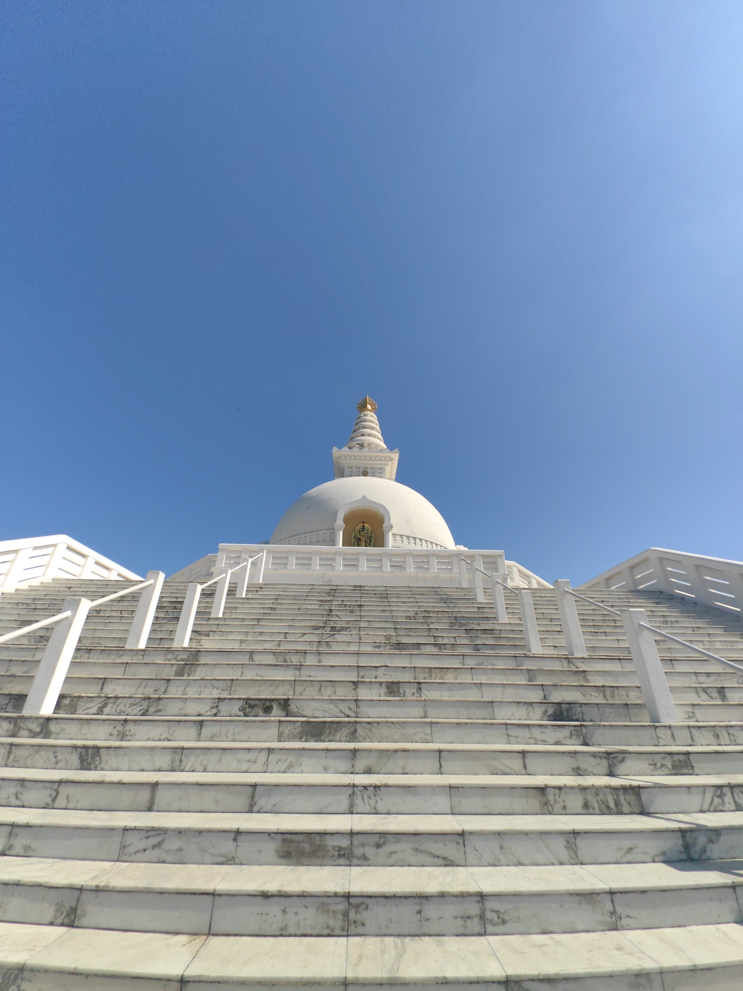 The image depicts the World Peace Pagoda in Lumbini, Parsa, Nepal. The structure is predominantly white with a golden spire. A wide, marble staircase leads up to the pagoda, flanked by white railings. The sky is a clear, bright blue. There are no people visible in the frame. The foreground is dominated by the steps, with the pagoda rising in the background. The architectural style is characteristic of a Buddhist stupa.