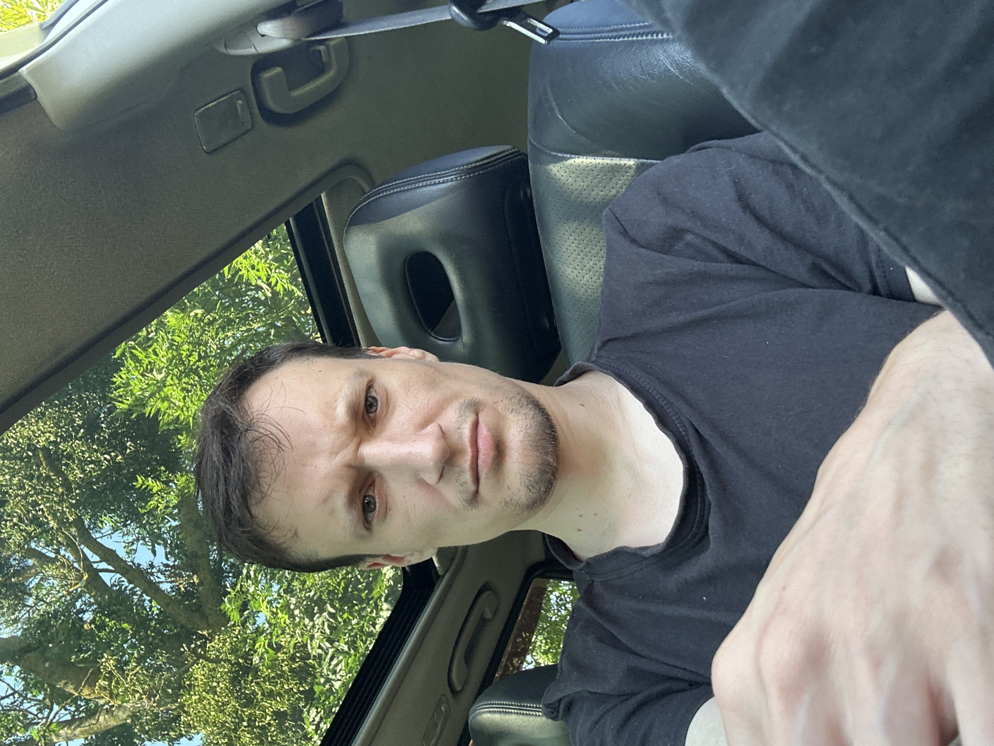 A medium shot photograph shows a man with a concerned expression sitting in the driver's seat of a car. He is wearing a black t-shirt and his left hand is visible resting on his leg. The car's interior is dark, with black leather seats and a sunroof open to reveal trees and a blue sky. The man's facial features are clearly visible, with furrowed brows and a slight grimace. The scene is likely captured on a sunny day, as indicated by the bright sunlight filtering through the trees and sunroof.
