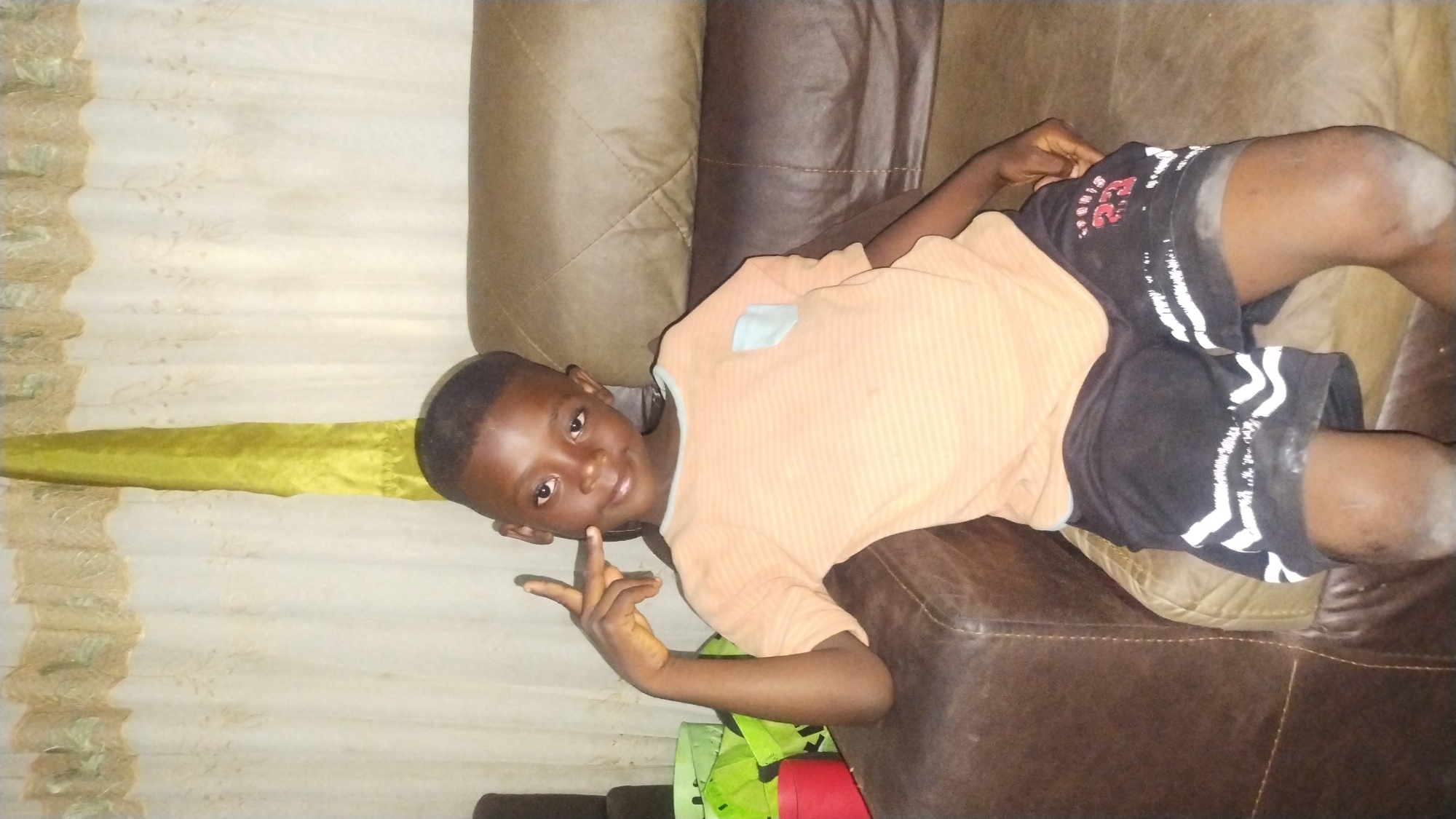 This image, captured in the city of Jos, Nigeria, depicts a single male child seated on a brown leather or faux leather sofa within an indoor setting. The child, estimated to be between 5 and 8 years old, has dark skin, short dark hair, and is looking directly at the camera with a slight smile or smirk.
He is wearing a light peach-colored, horizontally striped short-sleeved t-shirt with a light blue patch pocket on the left chest and light blue trim around the collar. His lower attire consists of dark gray or black athletic shorts featuring white patterns or stripes on the lower legs and the red text "SPORTS 23" on the right thigh. A white powdery residue is visible on his knees and lower thighs.
The child's left arm is bent, with his hand positioned near his left cheek, forming a "V" sign with his extended index and middle fingers. His right arm rests on the armrest of the sofa.
The sofa features visible stitching and quilted sections on the backrest and armrests. To the immediate left of the child, on what appears to be an adjacent surface, are stacked items including at least two bright green, possibly fabric, bags or containers, and a red circular container. The background consists of light-colored, subtly patterned curtains, partially obscured by a darker patterned valance. A vertical strip of bright yellow or lime green fabric hangs down to the left of the child's head, likely a curtain tie-back. The scene is illuminated by an unseen light source, indicating an indoor domestic environment.