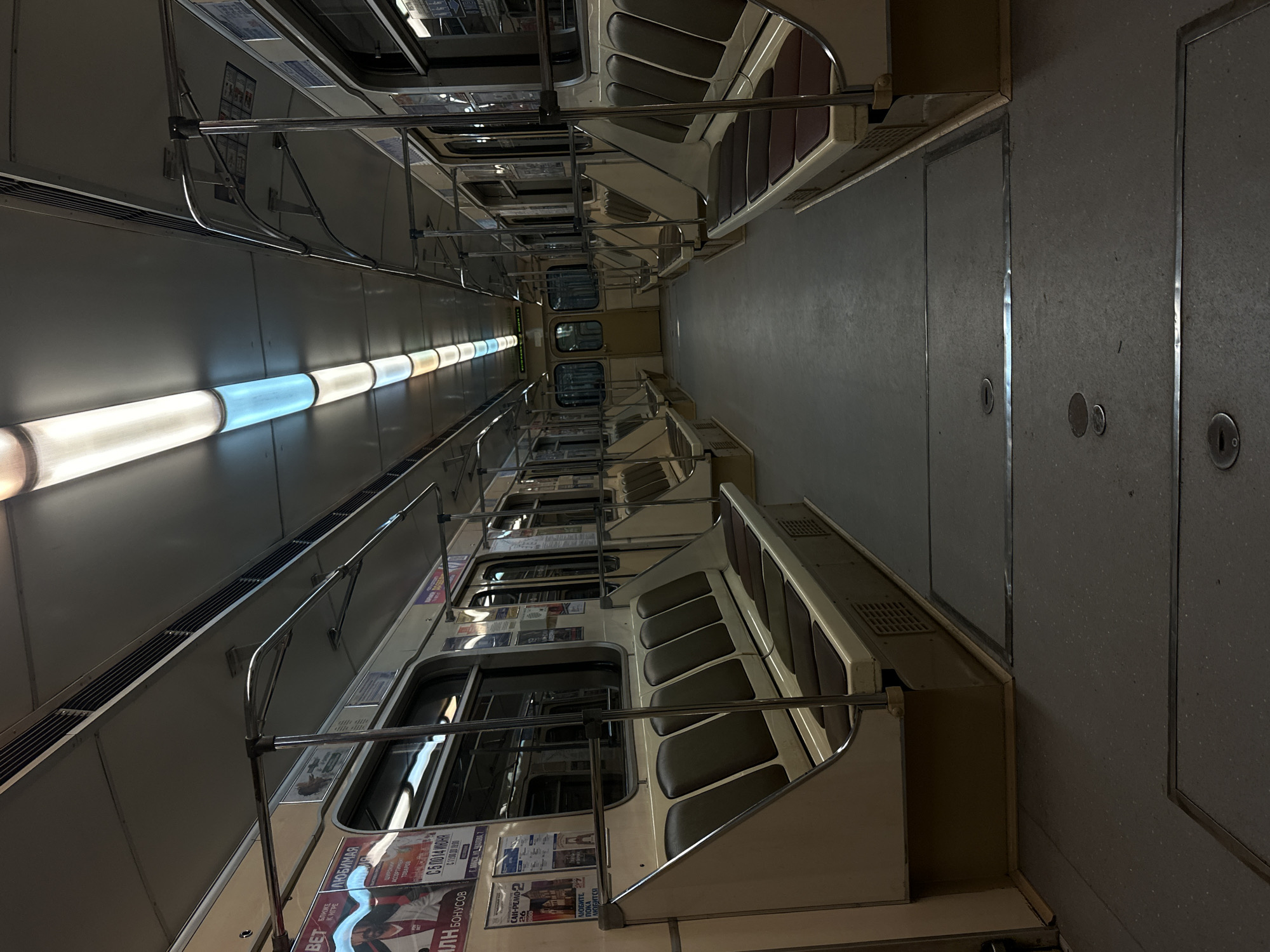 The image shows the interior of a subway car in Minsk, Belarus. The car is empty of passengers. It features rows of beige seats with dark brown cushions on either side of a central aisle. Stainless steel handrails run along the ceiling and vertical poles are spaced throughout the car. The ceiling has linear fluorescent lights, some of which emit a bluish hue. Advertising posters are visible on the interior walls. The floor is a dark grey material with metal grates and access panels. The overall scene depicts a typical subway car in a state of disuse or between journeys.