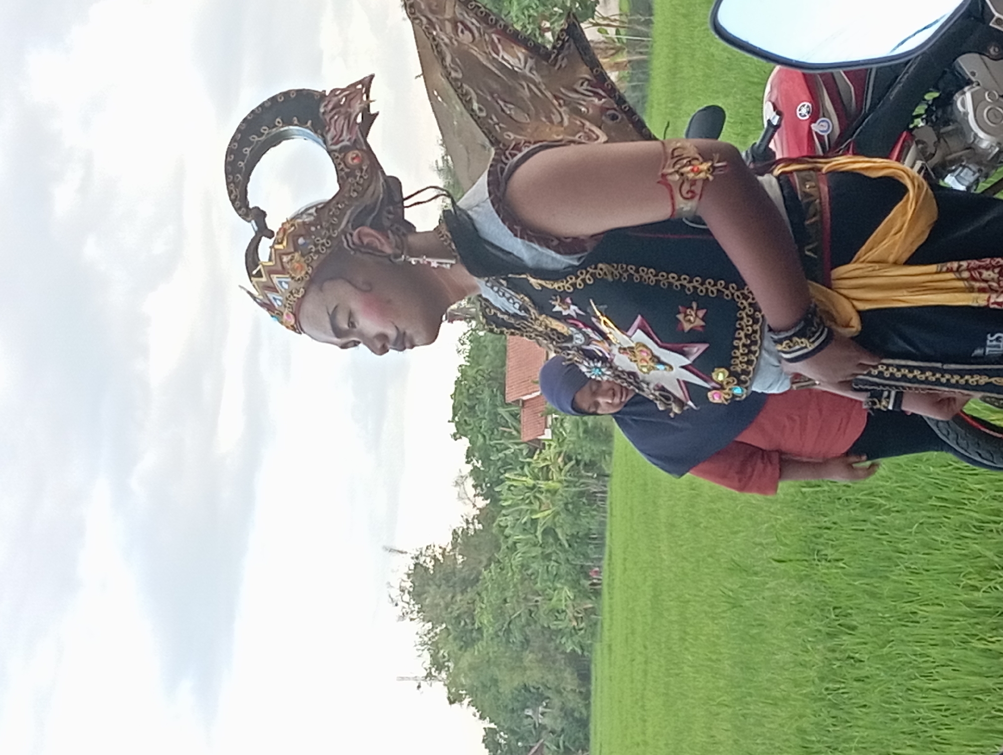 A person in traditional Javanese attire stands next to a motorcycle. The individual is wearing a headdress with a curved, ornate design and a black vest adorned with gold patterns. A yellow sash is draped over the vest. The person is positioned in front of a rice paddy field. In the background, there are trees, and a second individual is partially visible. The scene appears to be in a rural setting in Waung, Indonesia.