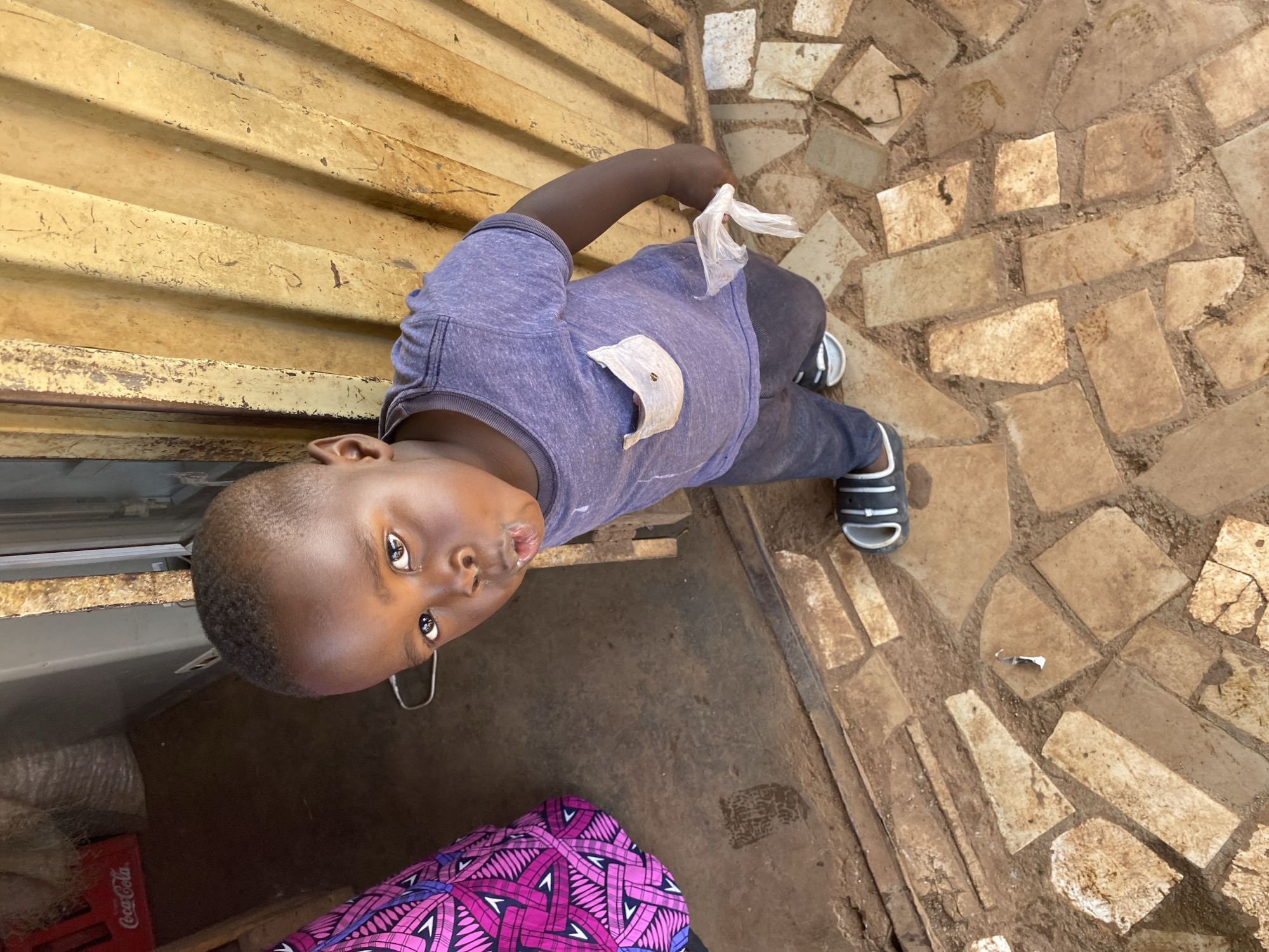 An overhead shot captures a young child, likely a boy, looking up towards the camera. The child, with dark skin and a close-cropped haircut, is centered in the frame. He wears a faded purple or grey t-shirt with a light-colored, possibly worn, patch on the left side of the chest, dark trousers, and black and white striped sandals. His right hand holds a crumpled clear plastic bag. A white earbud cable hangs near his neck.
The setting appears to be outdoors, in what could be a marketplace or the front of a small shop in Bukuru, Nigeria. Behind the child, a corrugated metal wall, painted in a faded yellow-brown, acts as a backdrop. The ground is paved with irregularly shaped stone or concrete slabs, creating a rustic, somewhat worn pattern. To the lower left of the frame, a red Coca-Cola crate is partially visible, containing what appears to be bottles or plastic bags. The child's expression is neutral, with wide, dark eyes looking directly upwards. The lighting suggests it is daytime.
Visible text includes "Coca-Cola" on the red crate. The overall scene depicts a casual moment in an everyday, possibly commercial, environment.