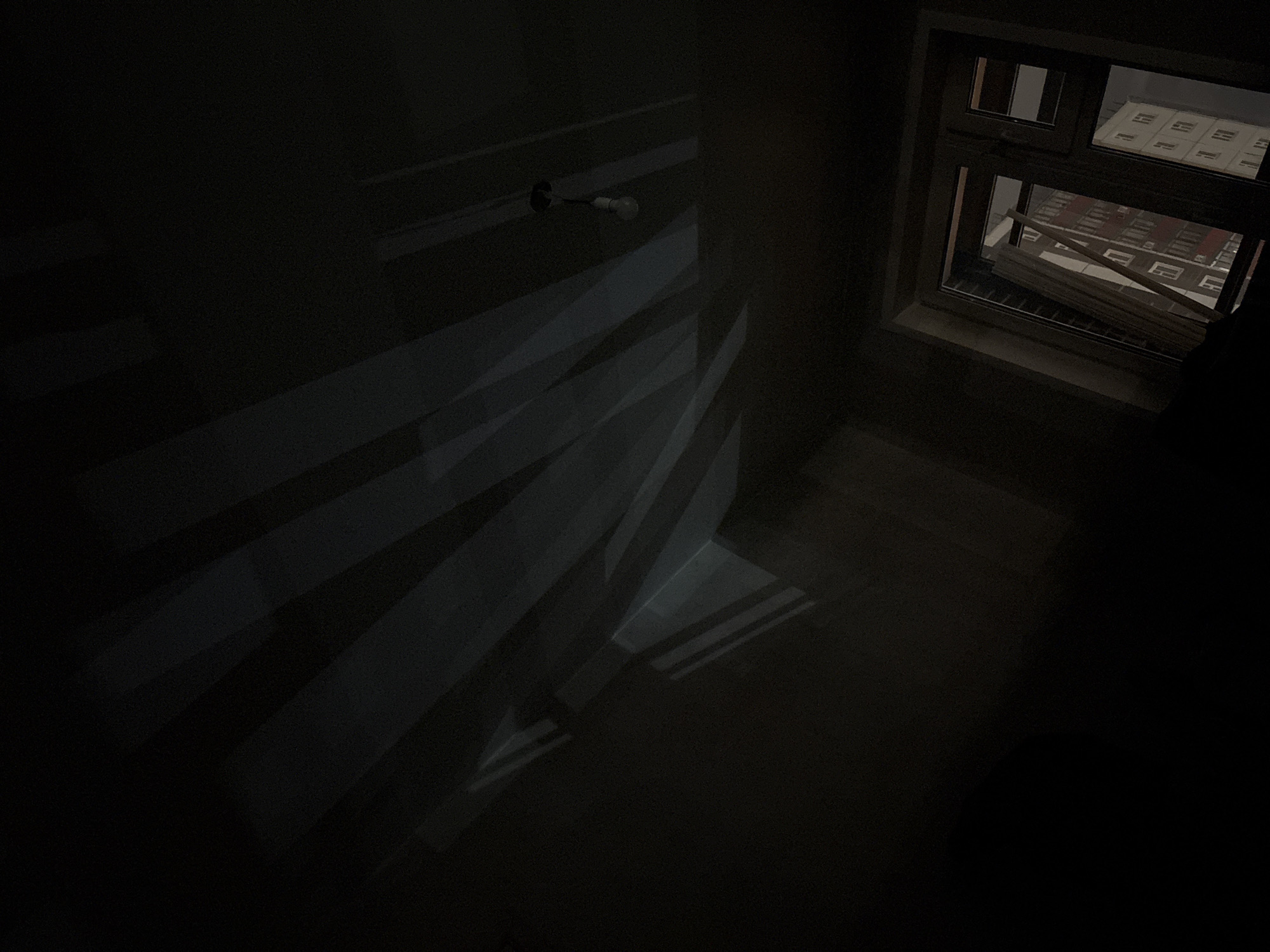 A dimly lit, interior view reveals a room under construction. Bare walls are visible, with a light bulb fixture extending from the wall. Strips of light and shadow pattern the left wall. A window on the right allows light to enter, and a partial view of the cityscape outside can be seen. Based on the visible architecture of the building and street patterns visible outside the window, it is likely somewhere in Krasnoyarsk, Russia. The window is partially open, with something like wooden blinds or construction materials resting on the sill. The overall impression is that of a room in transition, possibly undergoing renovation or still under construction.