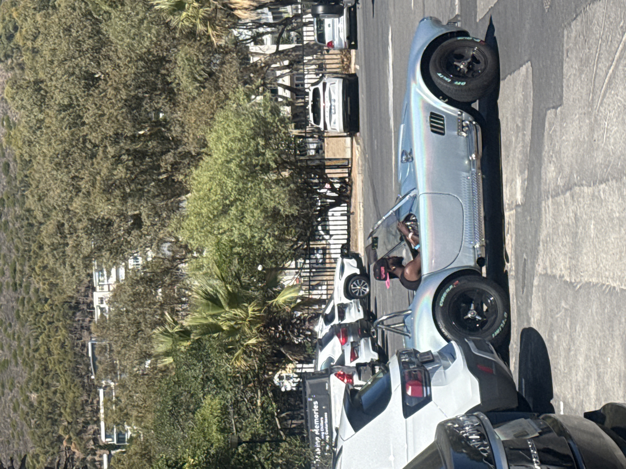 The image shows a silver sports car, possibly an AC Cobra replica, parked on a paved road in what appears to be a neighborhood setting. A person with a pink baseball cap is visible in the driver's seat. Several other cars are parked along the road. The background includes trees, a fence, and buildings that are indicative of a residential or mixed-use area. Based on the palm trees and building styles, it is probable that this scene is located in Cape Town, South Africa.