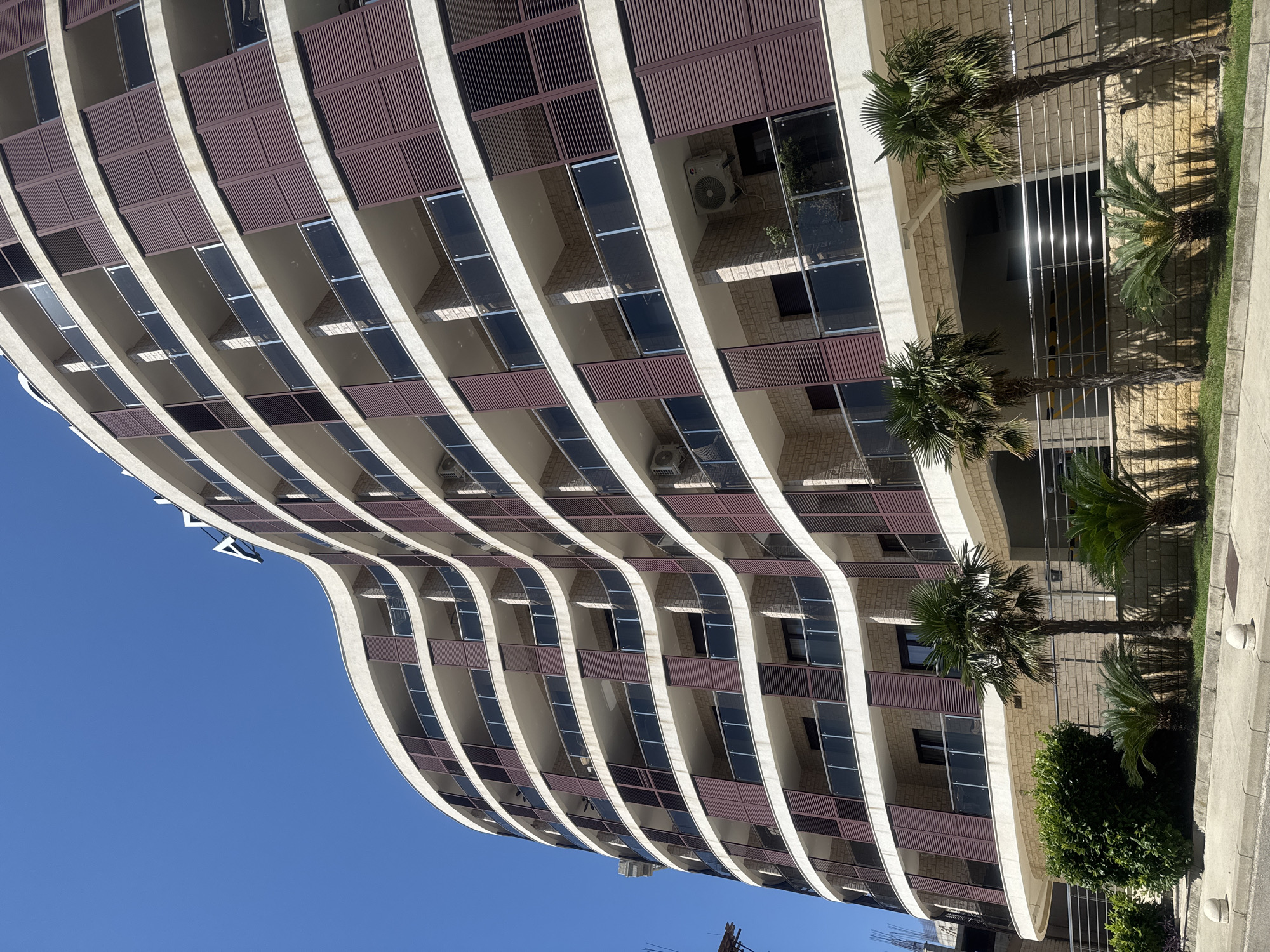 This is a low-angle, eye-level shot of a modern, curvilinear apartment building in Boreti, Montenegro. The building features multiple floors with balconies and dark red shutters. The facade is a mix of light-colored stone and white trim. Several palm trees and lush green bushes are situated in front of the building. The sky is a clear, bright blue. The ground in front of the building is paved with a sidewalk and street. There are no people visible in the image.