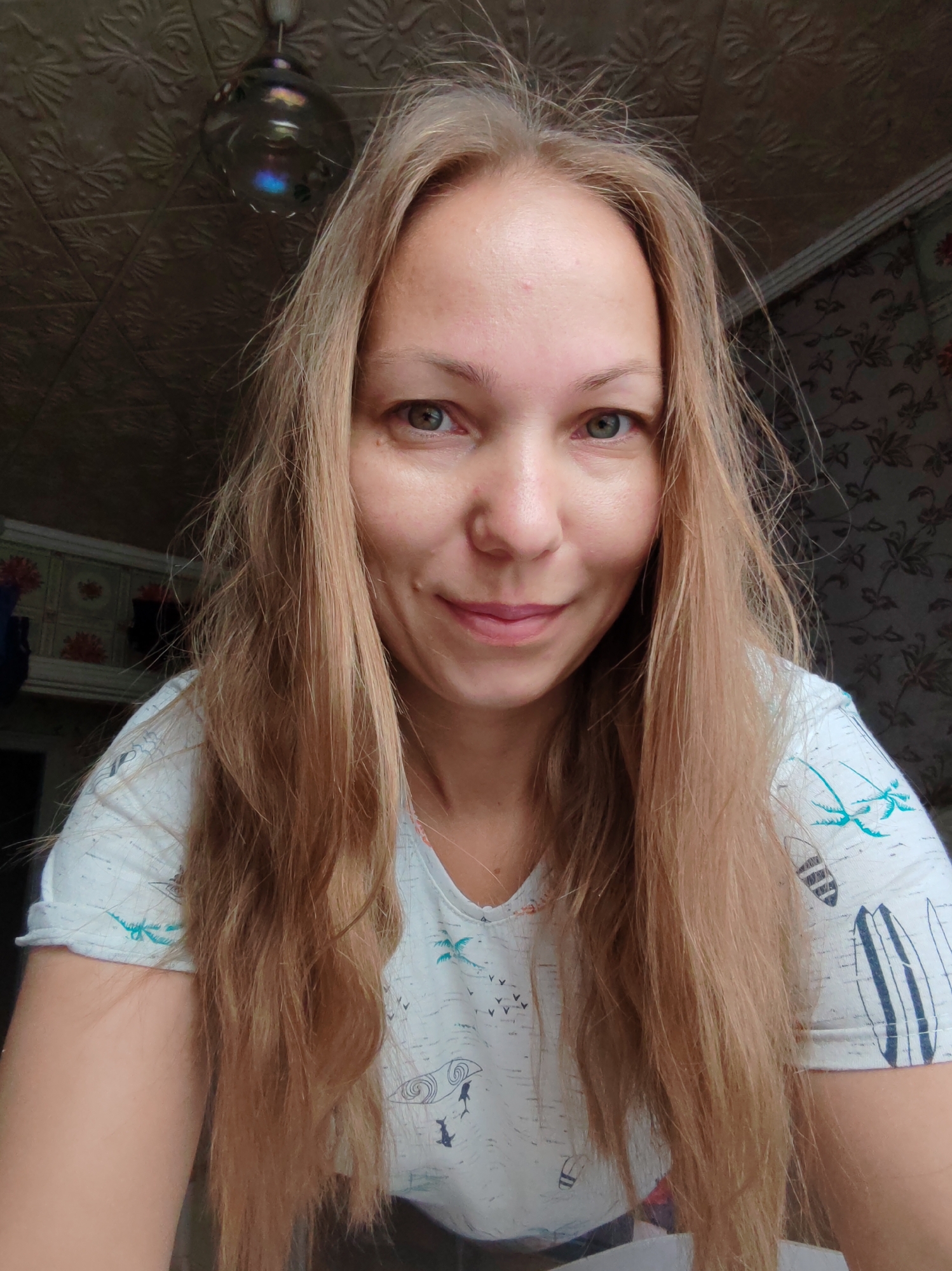 A medium shot shows a fair-skinned woman with long, straight blonde hair and light green eyes. She is looking directly at the camera with a slight smile. She is wearing a white t-shirt with a pattern of surfboards and palm trees. The background is a wall with a textured pattern and wallpaper with a floral design. There is a hanging lamp visible in the upper left corner of the image. The scene appears to be indoors, possibly a home, in the city of Chukhlem, Russia.