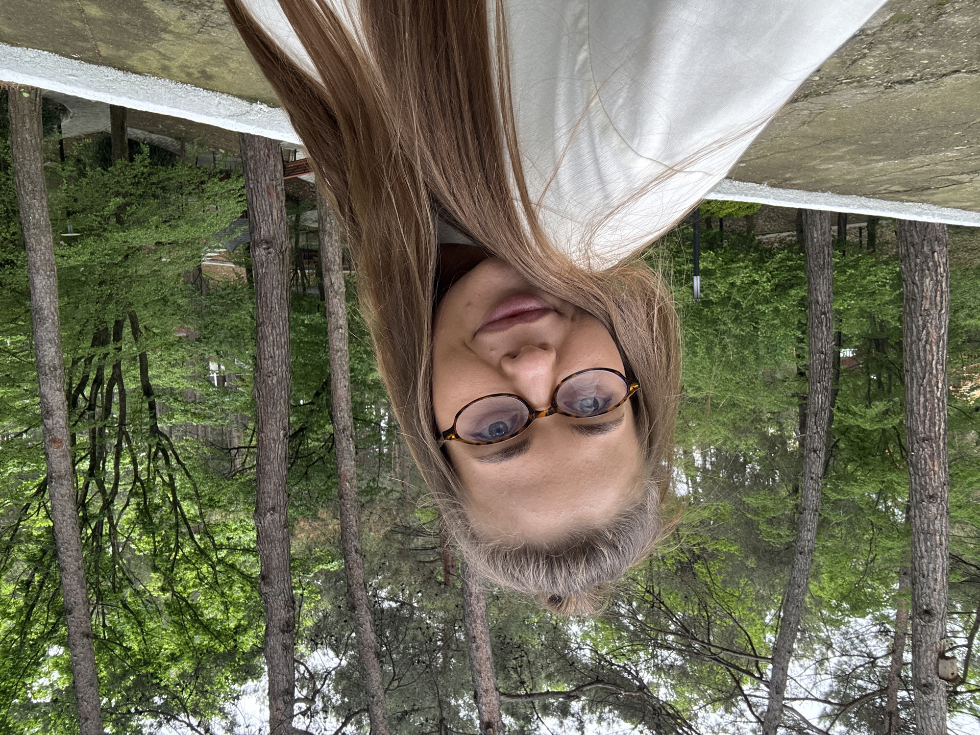 A woman with glasses and long brown hair is in the foreground, while a forest scene is in the background. The woman is wearing a white shirt. The forest features tall trees with green leaves. The picture appears to be taken in Sosnovy, Russia.