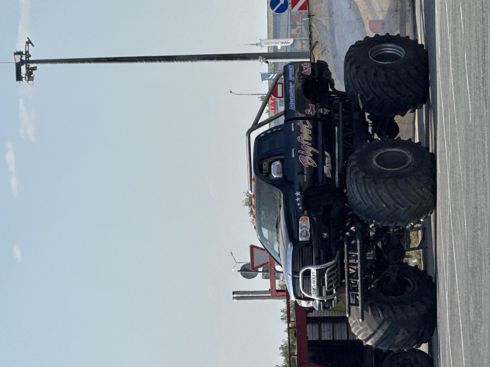 The image shows a black "Bigfoot" monster truck on a paved surface. The truck's tires are large and prominent. In the background, there is a street with signs and a tall metal pole. The location is Unknown, Unknown.