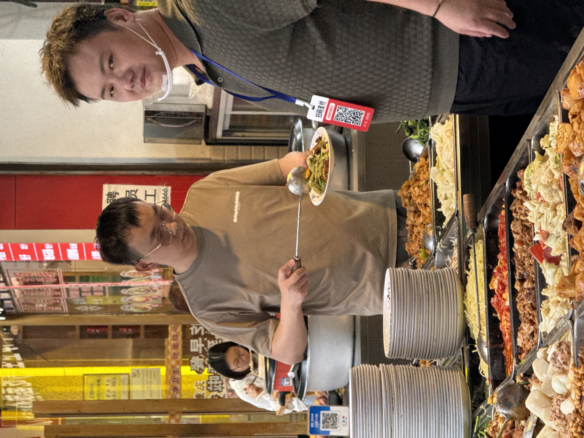 The image shows a man in a light brown t-shirt with the sleeves rolled up, holding a plate of food in his left hand and a serving spoon in his right. He appears to be working at a food stall or shop, evidenced by the trays of various cooked foods displayed to the right. Behind him, there are stacks of white plates.
Behind the man, another person can be partially seen in a white shirt. In the background, there are signs in Chinese, with one sign reading "聘员工" (Hire employees), and another sign indicating "干净卫生吃面放心" (Clean and hygienic, eat noodles with confidence), suggesting the location is a restaurant or eatery.
Above and slightly behind the man, another person is visible wearing a gray shirt and a clear face shield. They also wear a lanyard with an attached card that has a QR code and the Chinese characters "扫码支付" (Scan to pay).
Based on the OCR text "庆春早3 24666," the scene is likely in the Yuzhong District of China, specifically near a place called "Qingchun."