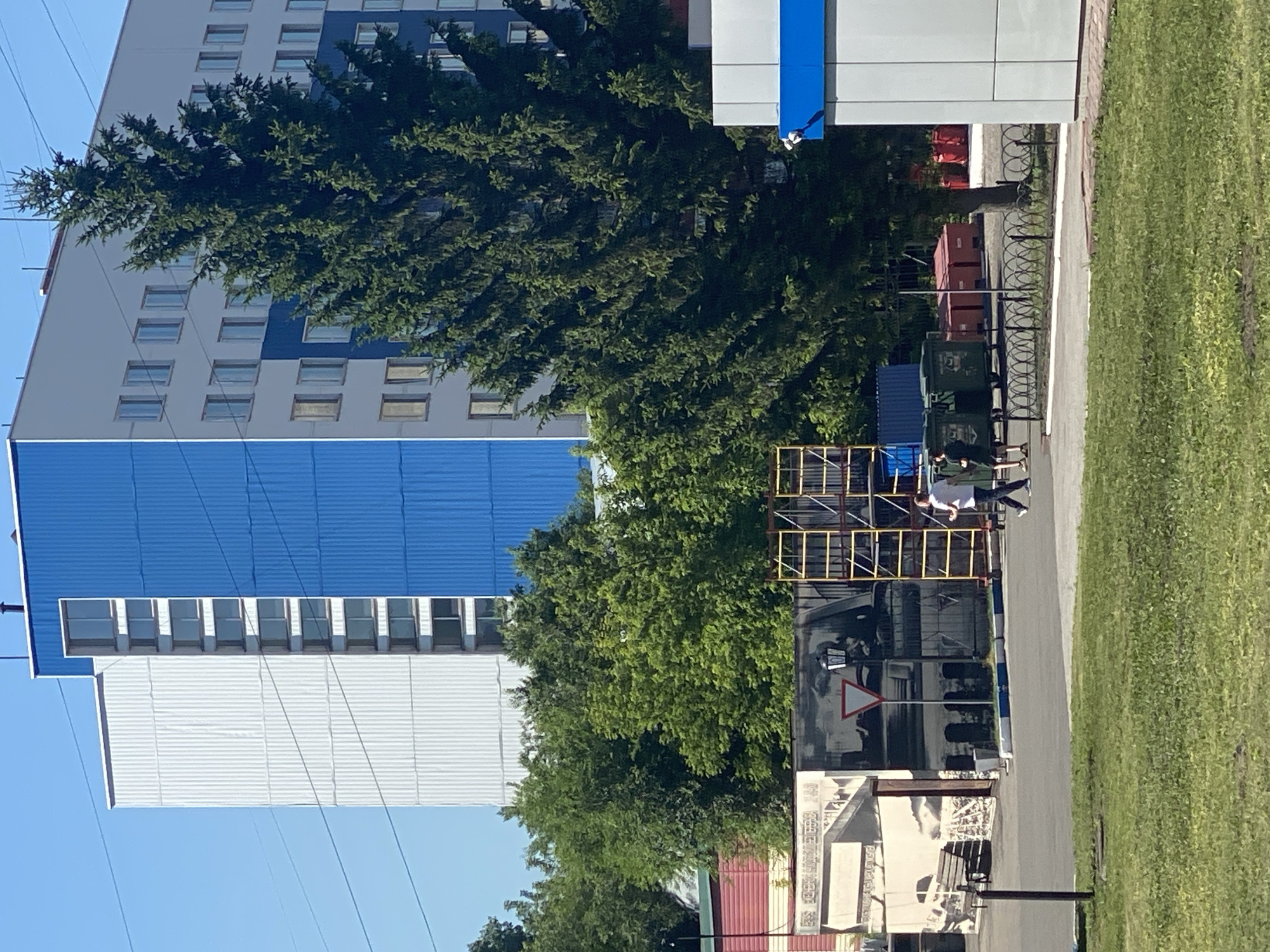The image depicts an outdoor scene in Novokuznetsk, Russia, on a clear day with a bright blue sky. A large, modern building with sections of white and bright blue paneling dominates the background. The building has numerous windows and appears to be multi-story. In front of the building, a tall evergreen tree stands prominently.
In the mid-ground, there is a mural or large advertisement on a wall, featuring black and white imagery. To the right of the mural, scaffolding is erected, and several large green dumpsters are visible, along with some red storage containers. Two individuals, one wearing a white shirt and dark pants, and the other in a dark shirt and pants, are walking across an asphalt area. A yield sign is posted in front of the mural.
The foreground consists of a grassy area and a paved path. In the far right, a portion of a white and blue building is visible. The overall impression is an urban or industrial setting.