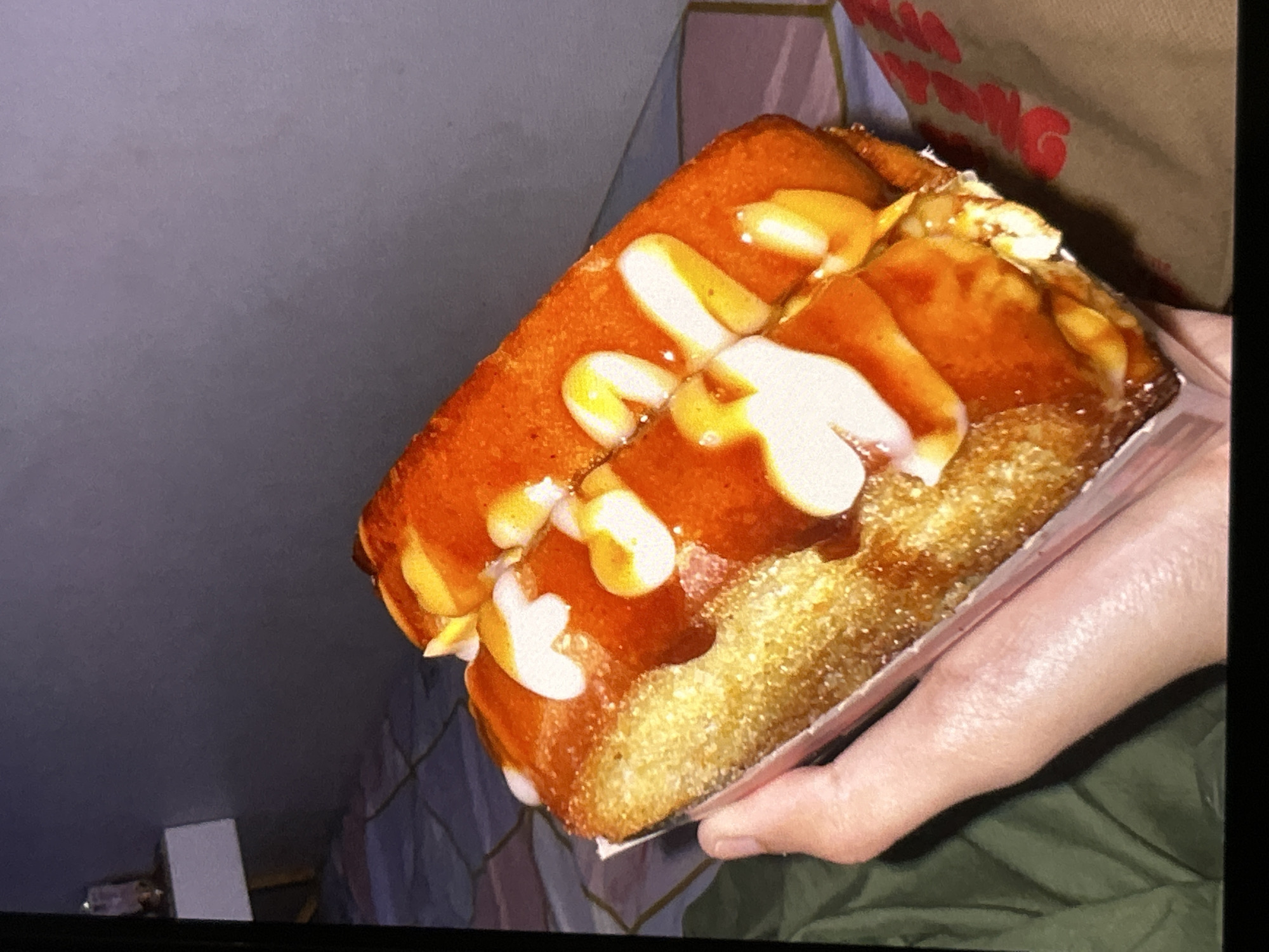 In Depok, Indonesia, a hand is holding a food item that appears to be a hot dog or corn dog, covered in a reddish sauce and white sauce. The background shows a bed with patterned sheets and a dark wall. The lighting is dim.