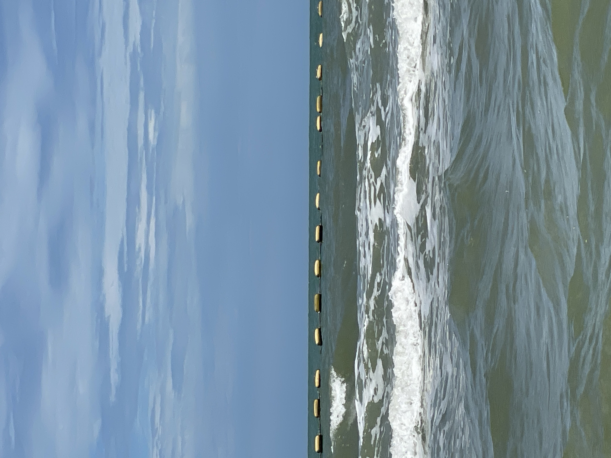 The image shows a view of the ocean with a line of yellow buoys stretching horizontally across the middle of the frame. The sea is moderately choppy with white waves breaking. The sky is cloudy and blue. The scene is likely a beach in Pattaya City, Thailand, given the typical weather and common sight of buoys marking swimming areas. There are no people or boats visible in the image. The overall impression is one of a calm, overcast day at sea.