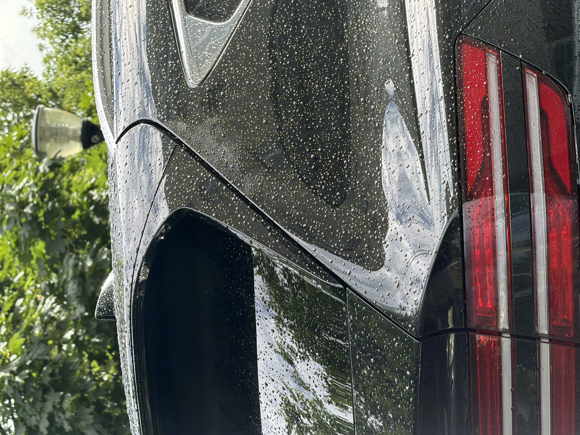 The image shows a close-up, side view of a wet, dark-colored car, likely in an outdoor setting with green foliage visible in the background. The car's surface is covered in numerous water droplets, indicating it has recently been rained on or washed. The focus is on the rear of the car, specifically the taillight assembly, which is comprised of red and white rectangular lights. Reflections of trees and the sky can be seen on the wet car panels, adding depth and detail to the scene. To the left, a portion of a street lamp is visible, partially obscured by the car and foliage. The lighting suggests it is daytime, possibly overcast or shortly after rain, given the water droplets and diffused light. The overall impression is one of a car exposed to the elements, with the water droplets creating a textured and reflective surface. There is no visible text in the image. The location context of Novaya Usman', Russia, places this scene in a specific geographic area, though the image itself doesn't contain explicit location markers.