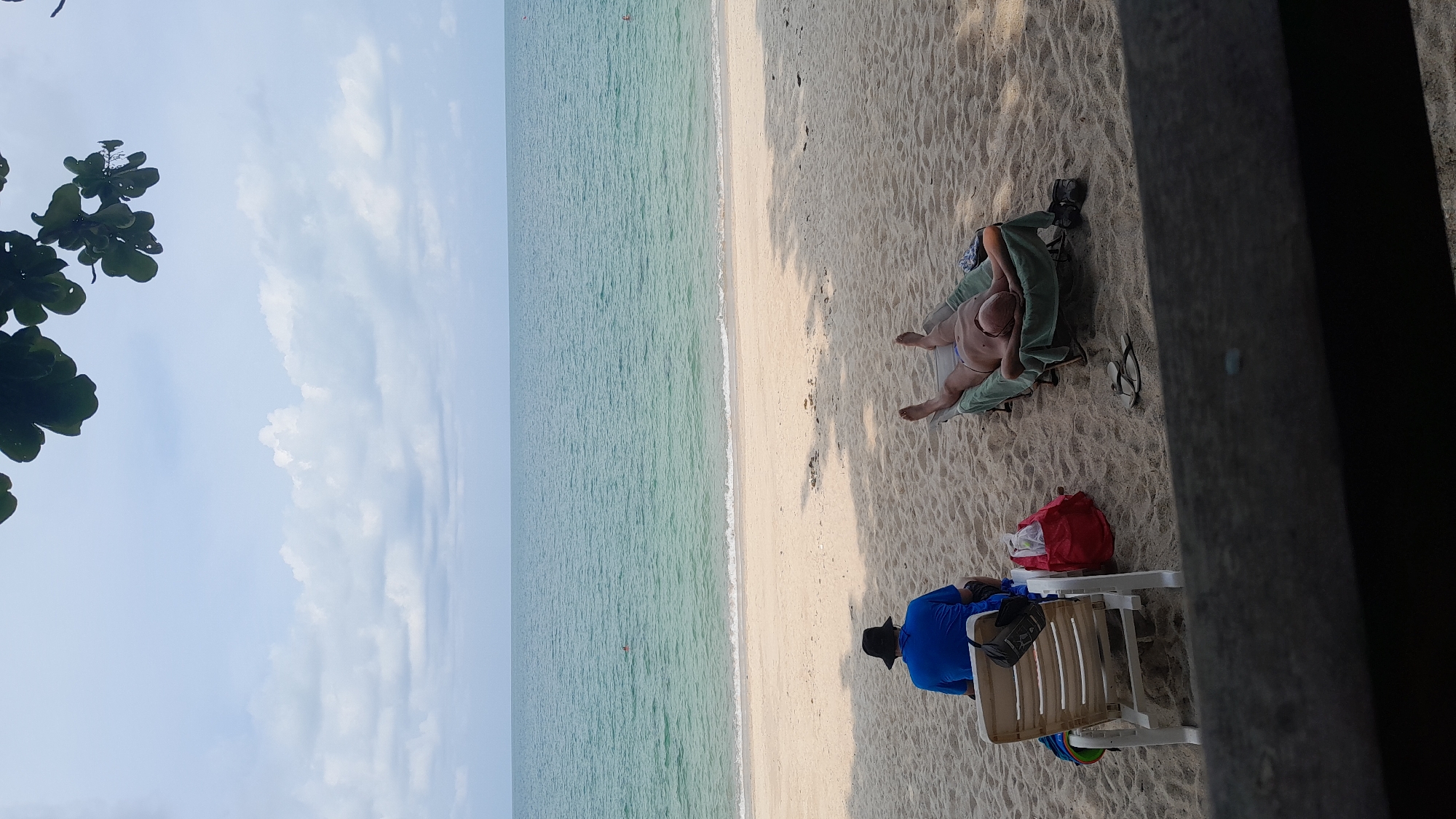 The scene is on a beach in Baan Hua Thanon, Thailand. The sea stretches to the horizon under a partially cloudy sky. On the sand, a man is reclined on a chair, covered in a light green cloth. Nearby, there are black shoes and flip-flops. A red bag sits on a small table next to a seated person in a blue shirt and black hat, who is occupying a white plastic chair. A tree branch is visible in the top left corner. A dark, blurred object is present on the right side of the frame.
