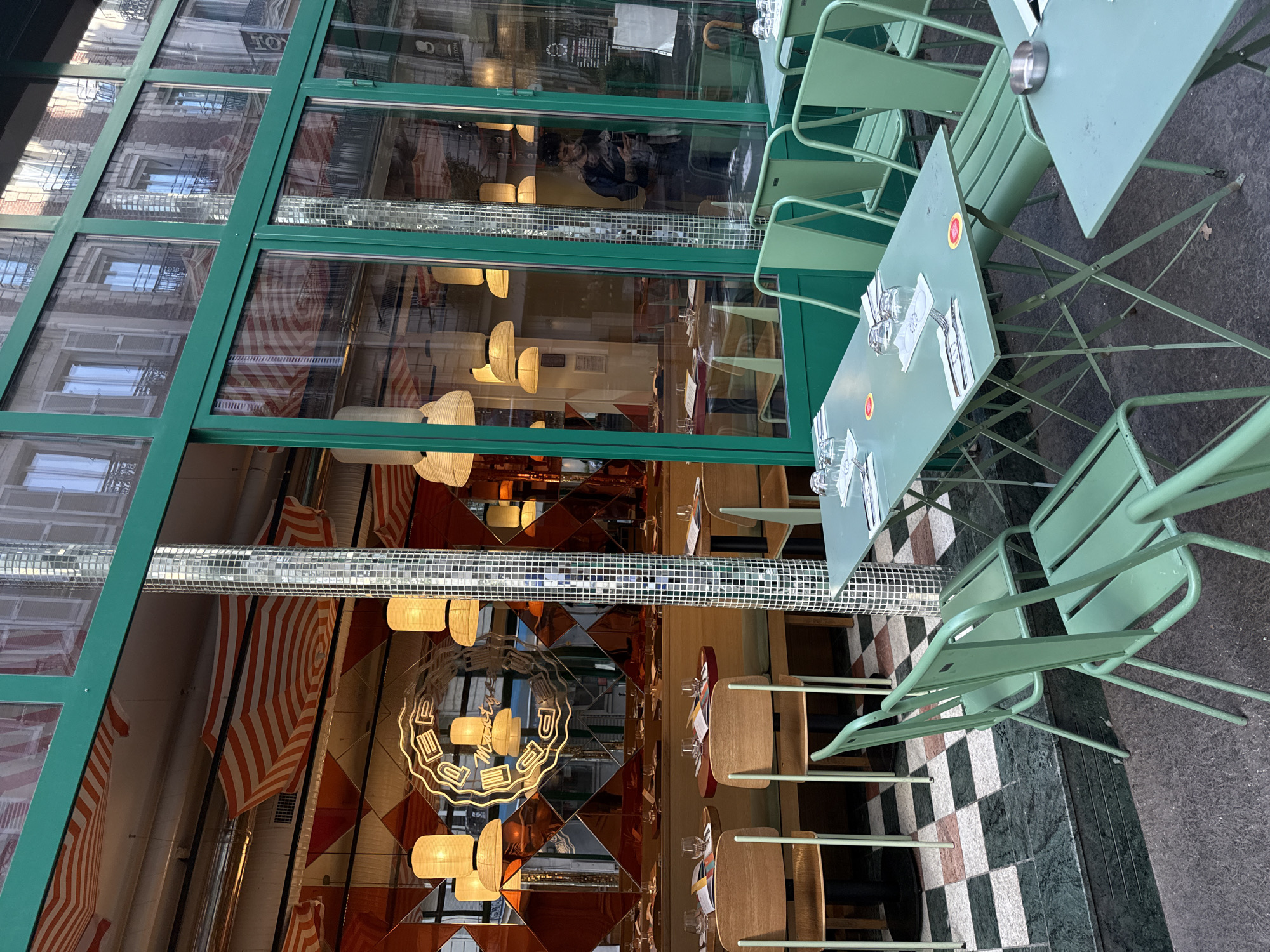 The media file, presented rotated 90 degrees counter-clockwise, depicts the exterior of a Parisian cafe with outdoor seating and a detailed view of its interior through large glass windows.
In the foreground, on the right side of the upright image, several light green metal cafe tables and chairs are arranged on a dark paved sidewalk. Some tables are set with white napkins, cutlery, and clear drinking glasses. Small red-bordered circular stickers, one clearly displaying "Coca-Cola," are visible on the tabletops.
The cafe's facade, located on the left side of the upright image, consists of expansive glass panels framed by dark green metal. Through these windows, the interior reveals a warm, eclectic decor. Walls feature orange and white striped awnings or panels, contrasting with sections of copper-toned, diamond-patterned mirrors. A prominent horizontal strip of small, reflective tiles, reminiscent of a disco ball, runs across the space. Warm light emanates from distinctive light-colored, possibly wicker-like, lampshades. A yellow neon sign is reflected in a mirrored surface, spelling "MAXI" and likely "PIZZA" or "PASTA." The visible indoor flooring is a checkered pattern of dark green, white, and reddish-brown tiles.
Reflected in the glass facade are the architectural details of traditional Parisian stone buildings across the street, complete with windows and iron balconies. A blurred reflection of a person in a dark coat and hat is also visible in the upper-middle glass panel. The lighting, with interior lights on and reflections of daylight, suggests either late afternoon or early evening. The scene is calm, with no active event or people directly observable, but the set tables indicate the cafe is open for business. The outdoor furniture style and reflected architecture strongly suggest the setting is Paris, France.